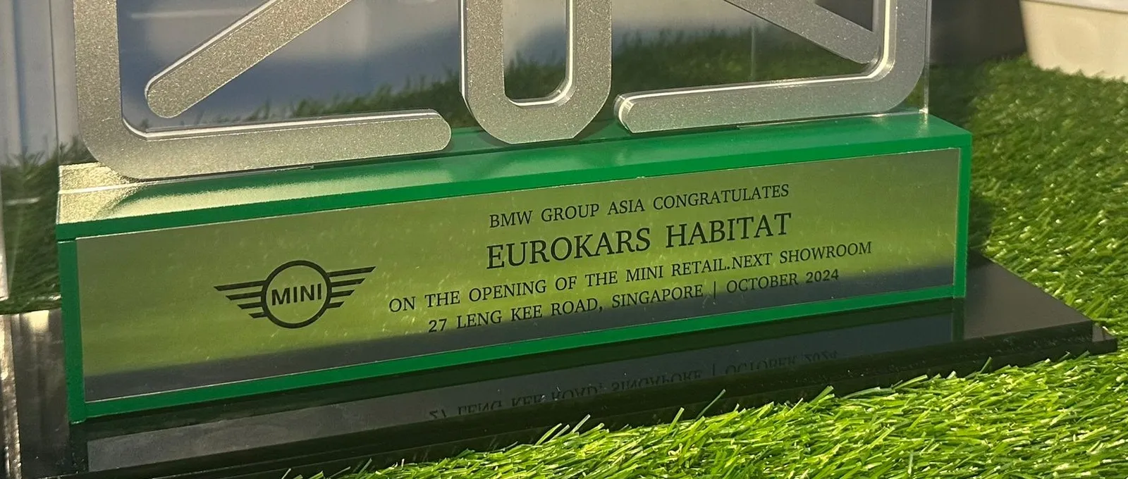 Close-up of the engraved plate on the green block: the MINI wings logo beside the opening message and showroom address, October 2024