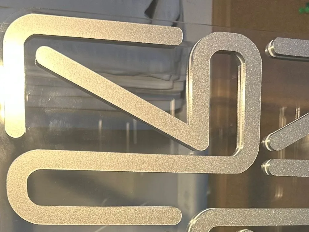 Close-up of the emblem's rounded strokes in fine-grained metallic silver, against the clear acrylic panel