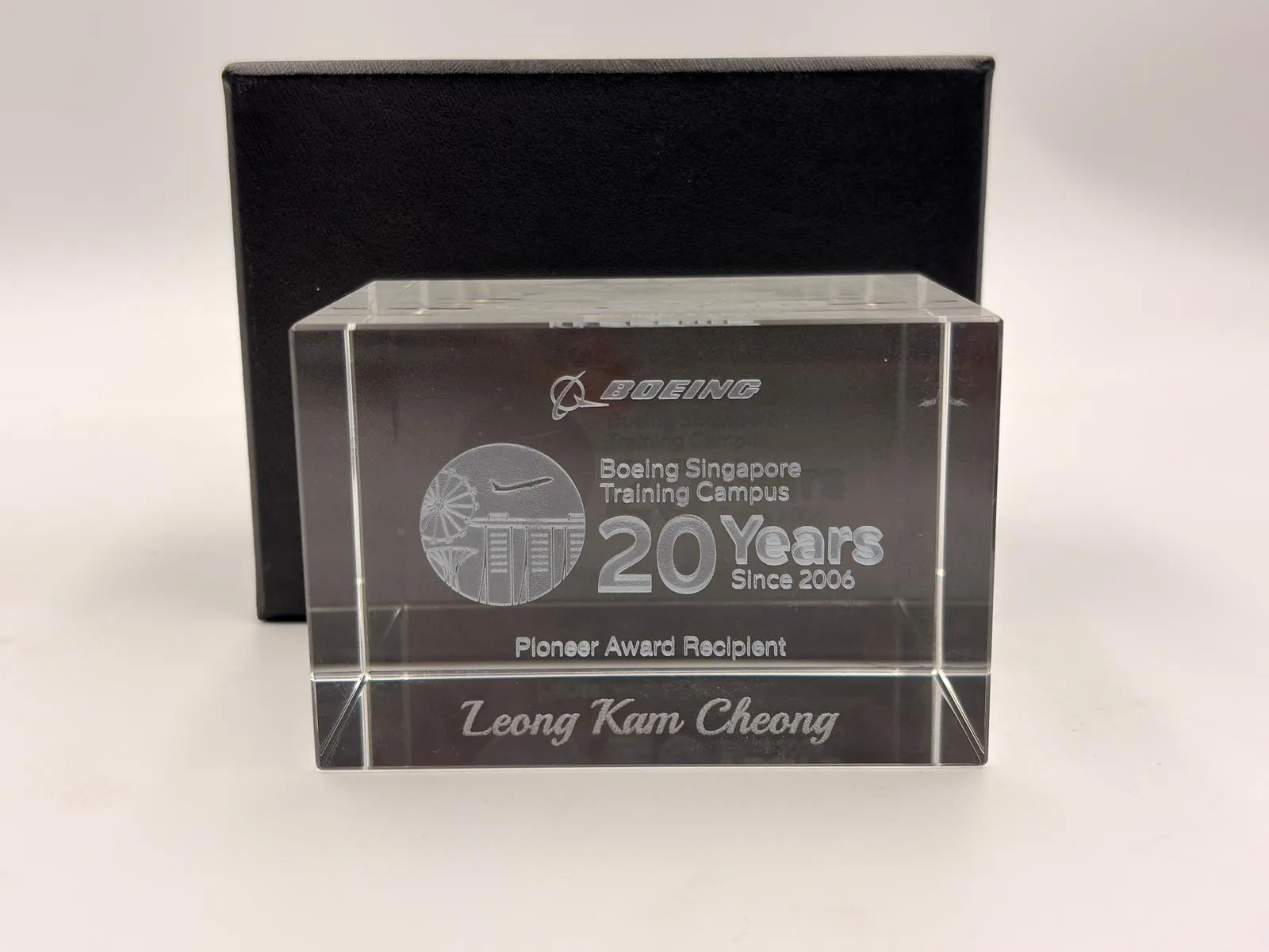 A clear crystal block engraved with the name Leong Kam Cheong, in front of a black box