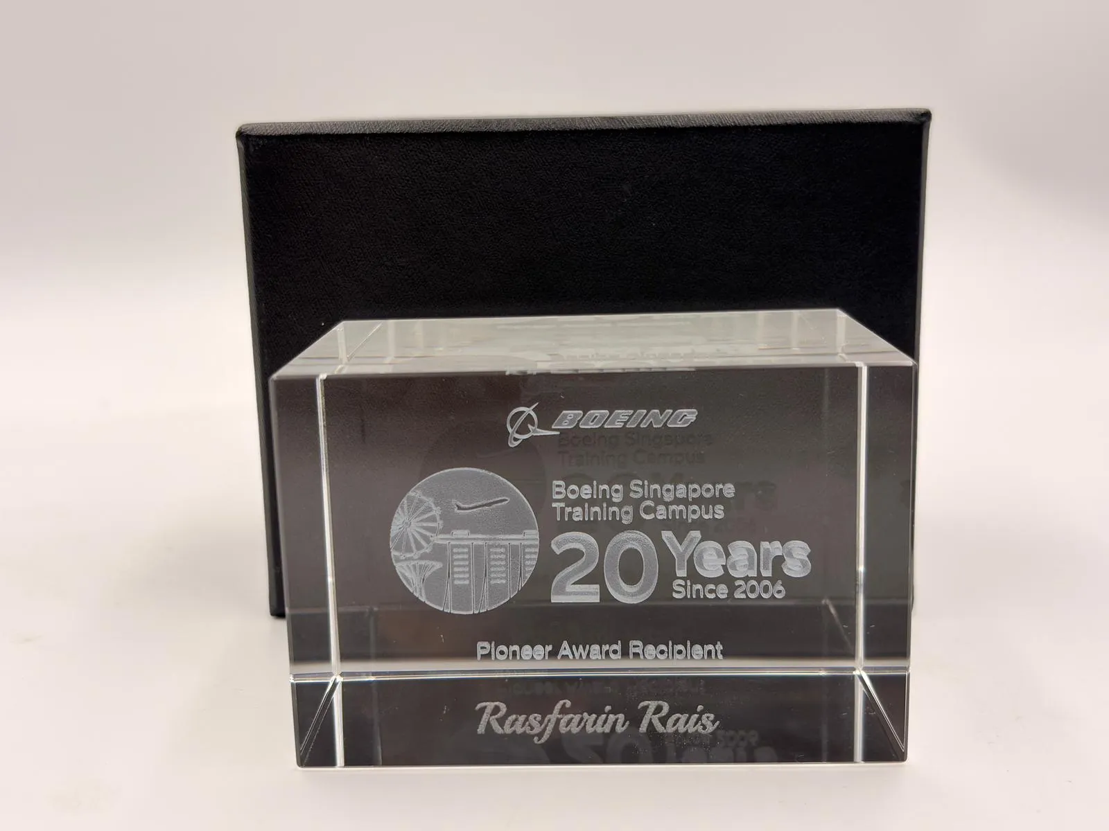 A clear crystal block engraved with the name Rasfarin Rais, in front of a black box