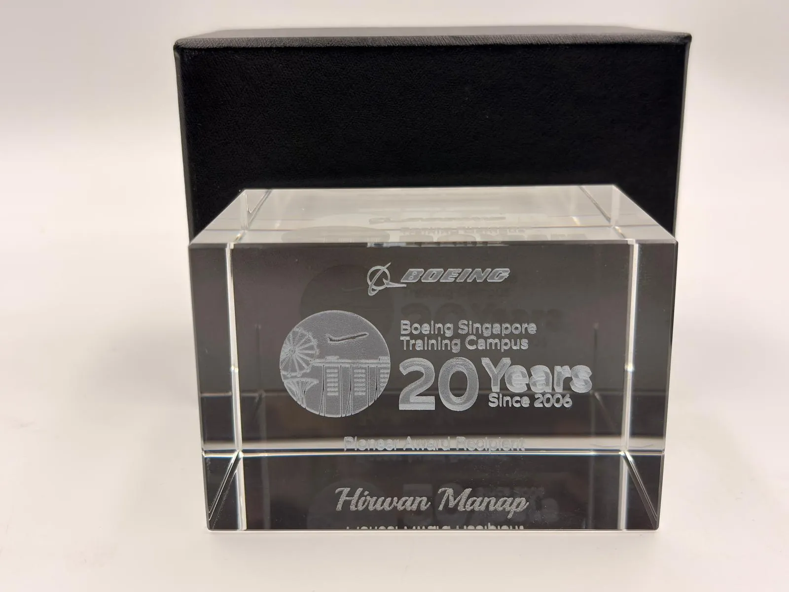 A clear crystal block engraved with the name Hirwan Manap, in front of a black box