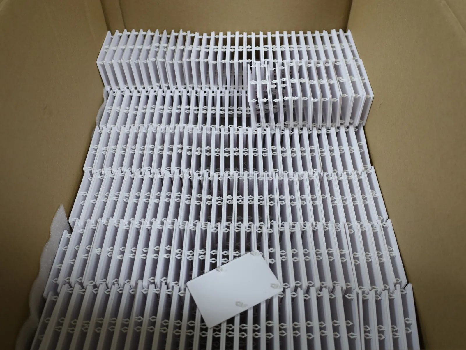 A carton packed with rows of white 3D printed fridge covers standing on edge, their arrow-shaped clips in line, one loose cover lying on top