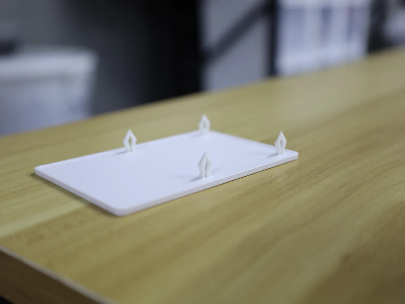 One white 3D printed fridge cover lying on a wooden desk, its four arrow-shaped clips pointing up from the flat panel