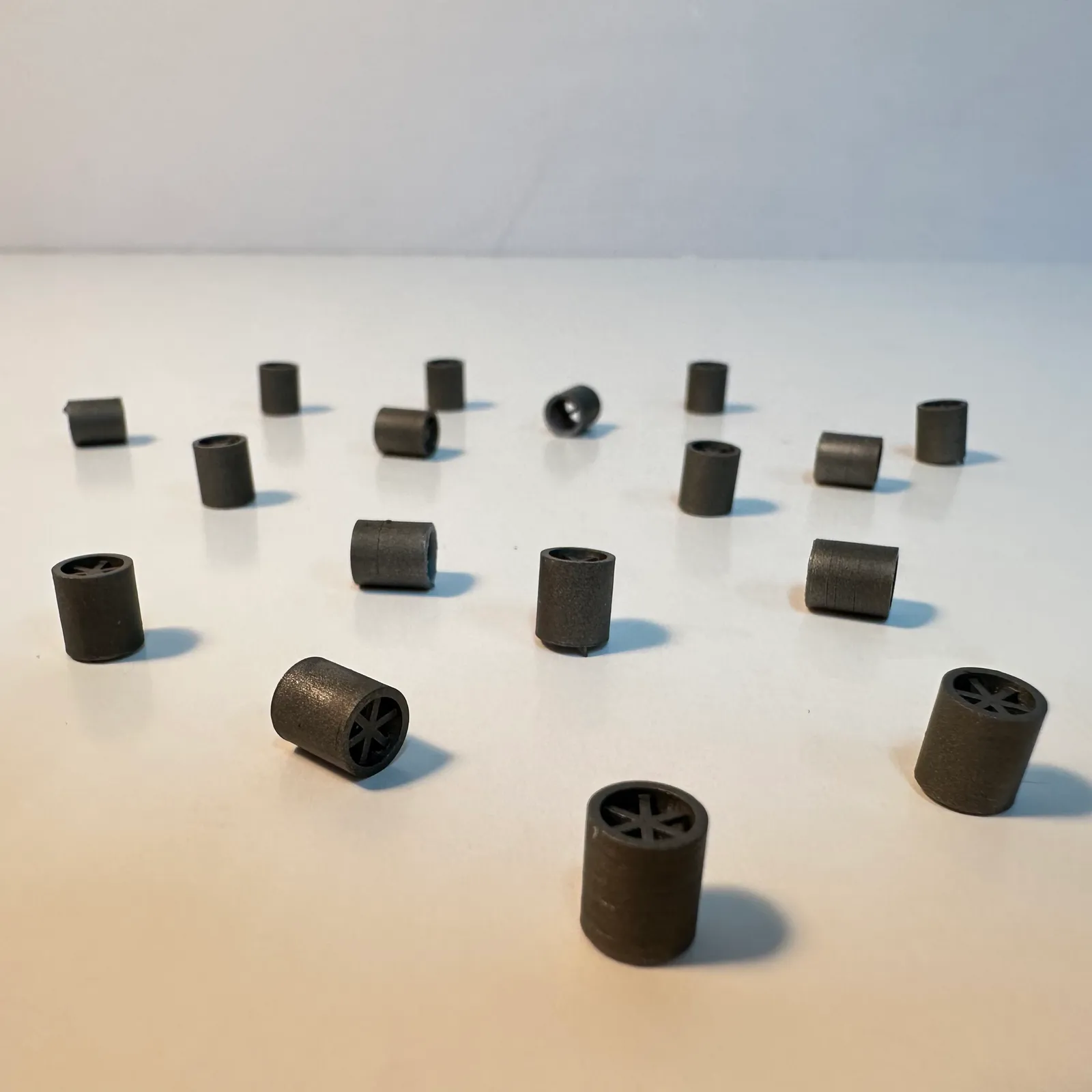 A batch of small black 3D printed spare parts spread across a white table, some standing upright and some on their sides, each a short cylinder with a six-spoke end