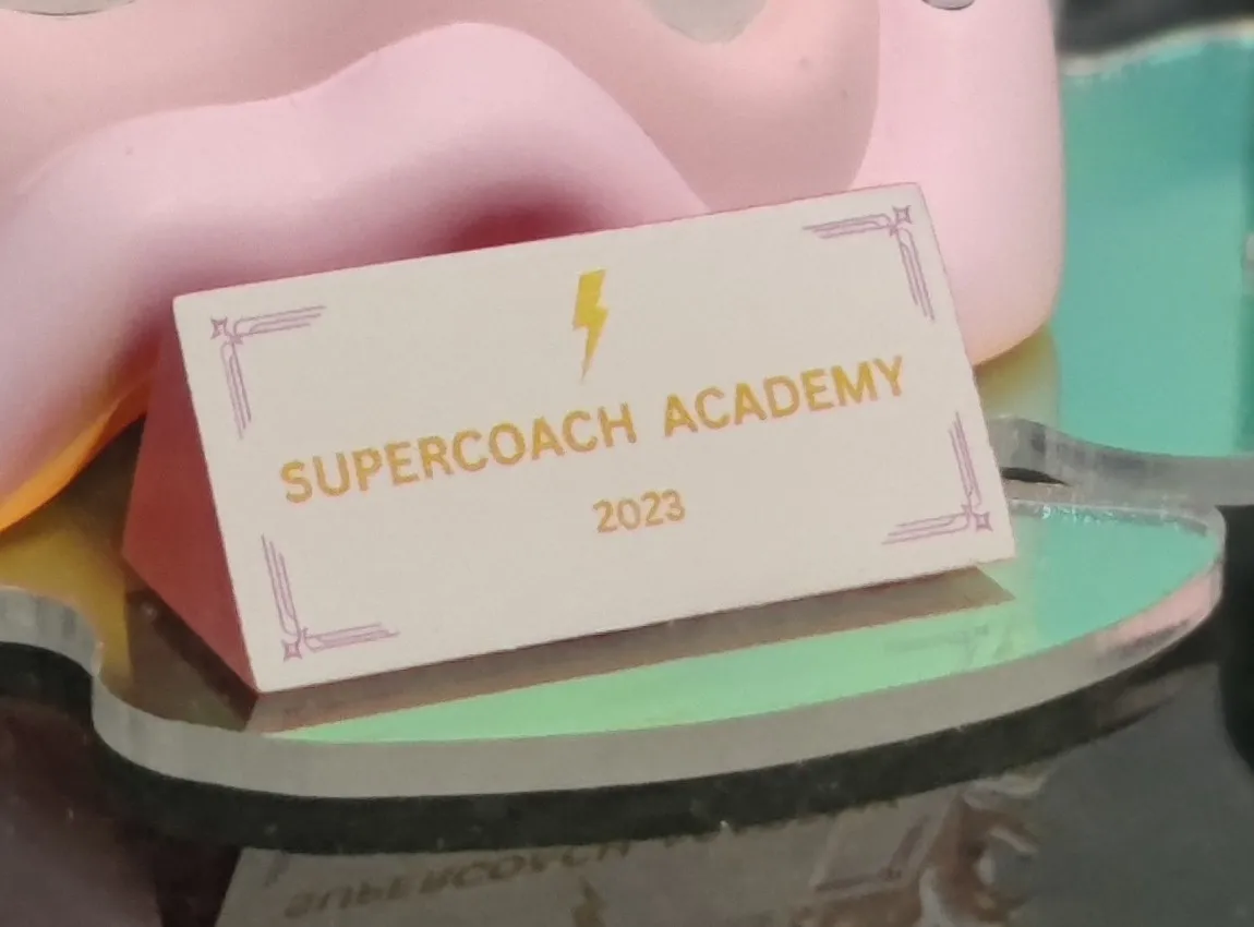 The cream tent-shaped nameplate with a gold lightning bolt and SUPERCOACH ACADEMY 2023 in gold, on the iridescent base