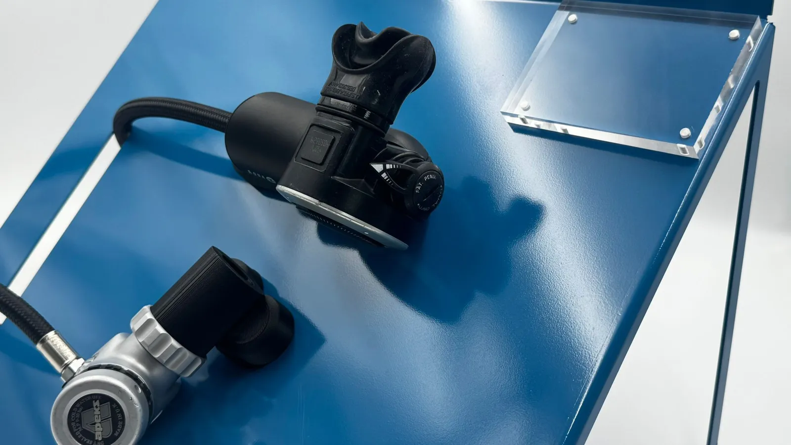 A regulator and spare mouthpiece held on the blue panel, the hose looping through the slot, beside the acrylic plate