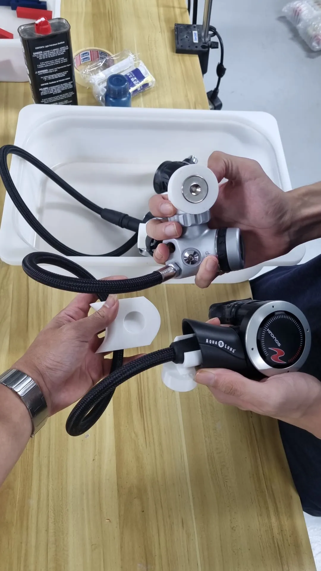 Two pairs of hands fitting white printed holders onto a regulator's first stage and second stage over a workbench