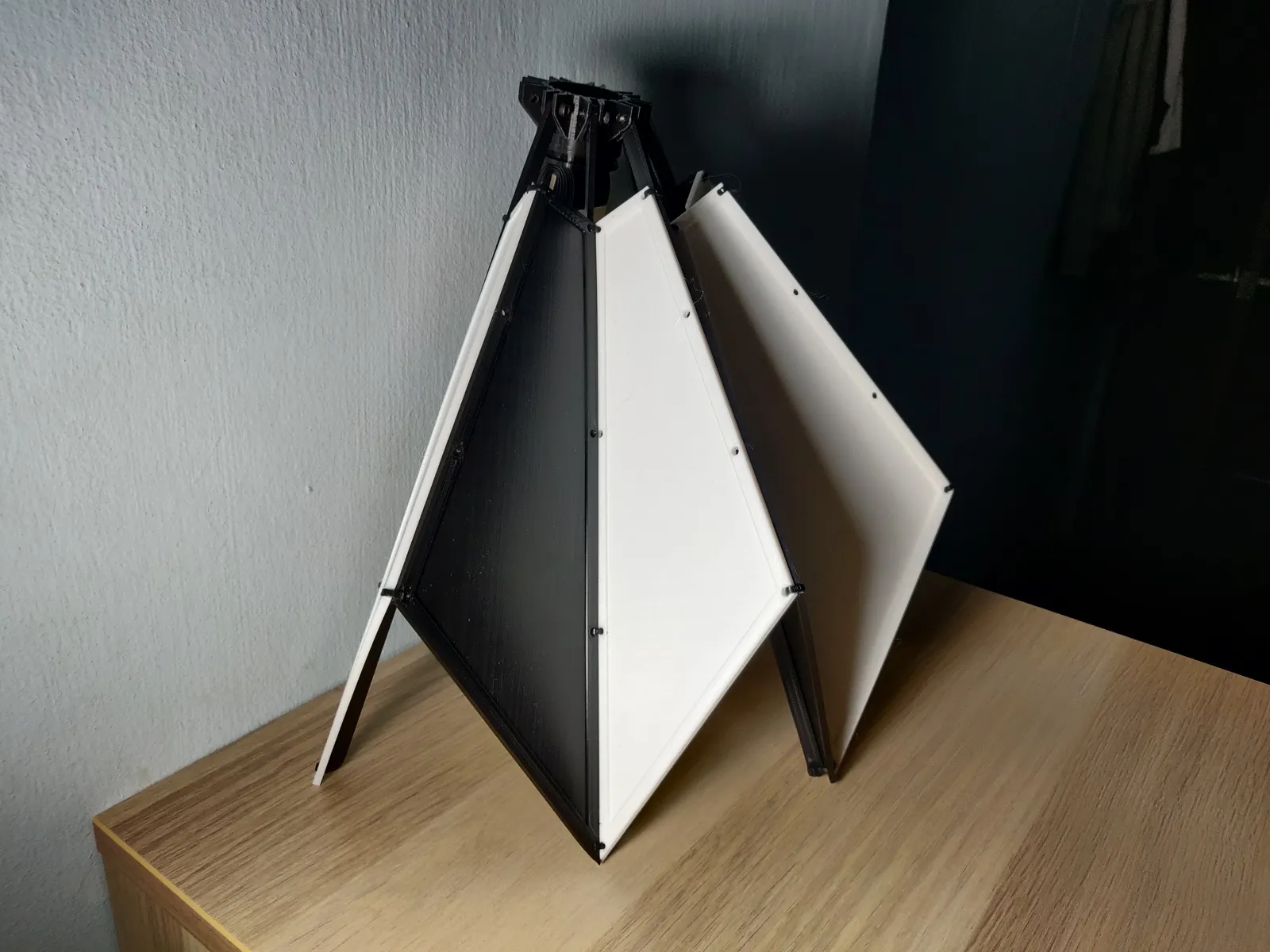The finished collapsible shelter prototype on a wooden desk: black and white 3D printed panels hanging from a black central hub, standing on their tips