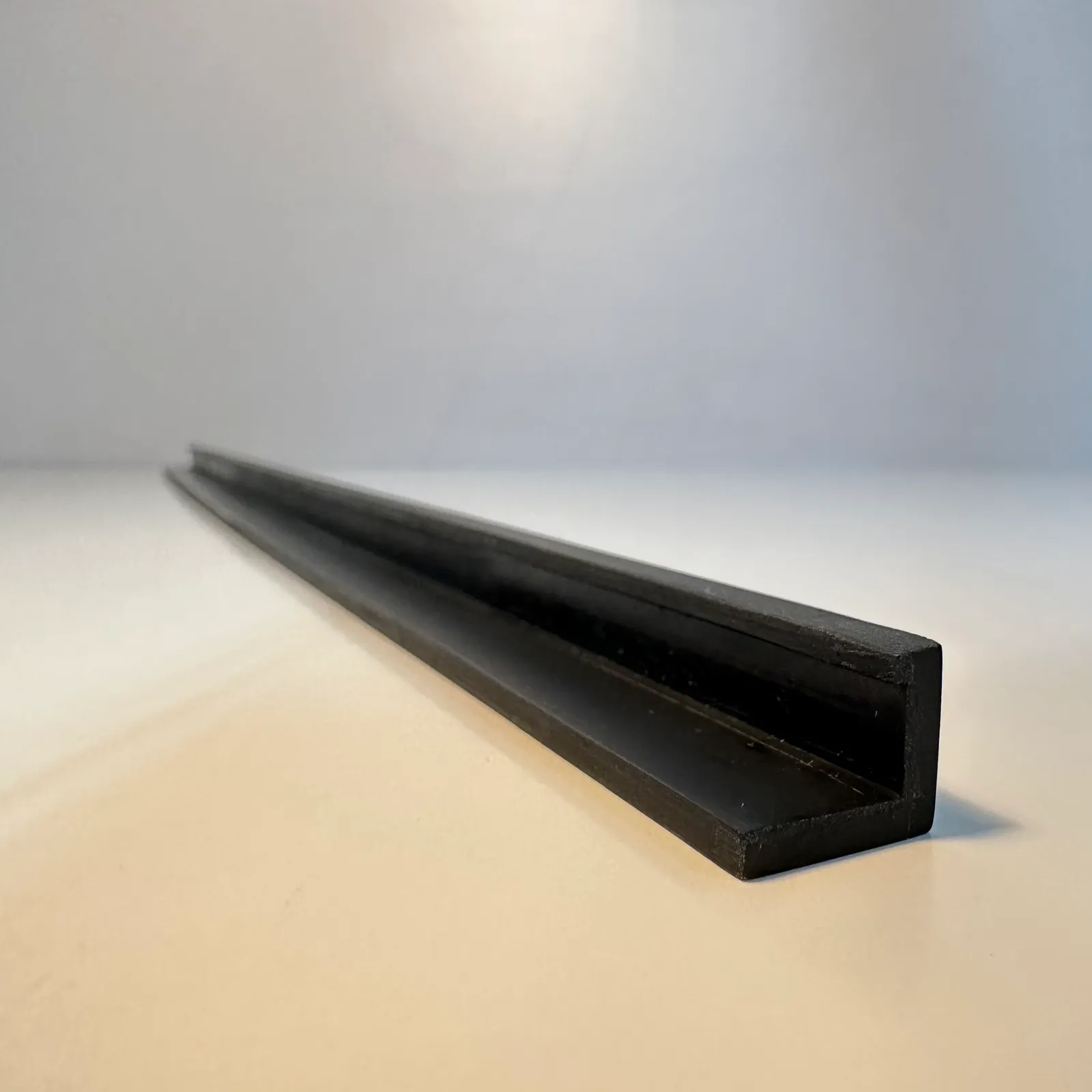The finished black angle trim strip on a white table, seen from one end: the right-angled profile, the lip along the top of the upright and the strip running away into the distance