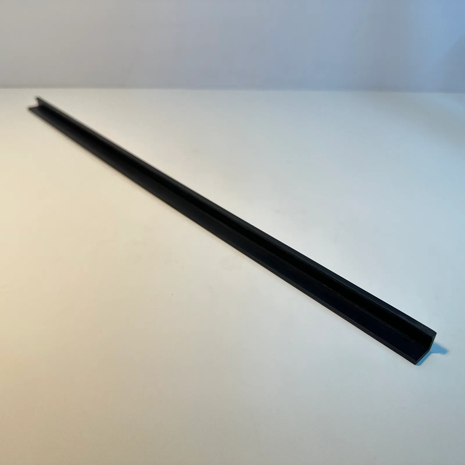 The whole black angle trim strip lying diagonally across a white table, seen from one end along its length