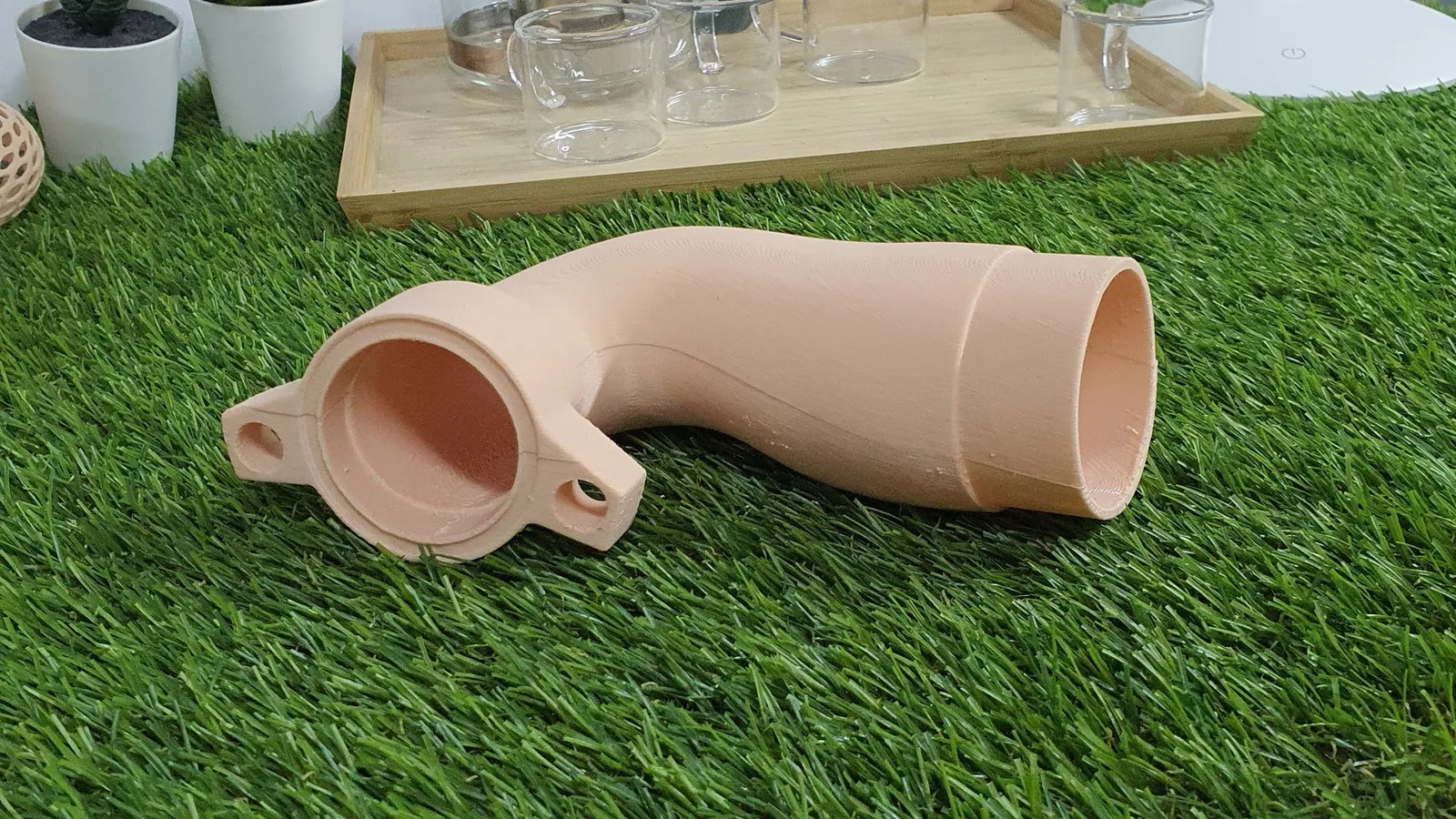 A 3D printed pipe elbow in beige lying on artificial grass, a round flange with two bolt holes at one end and an open socket at the other