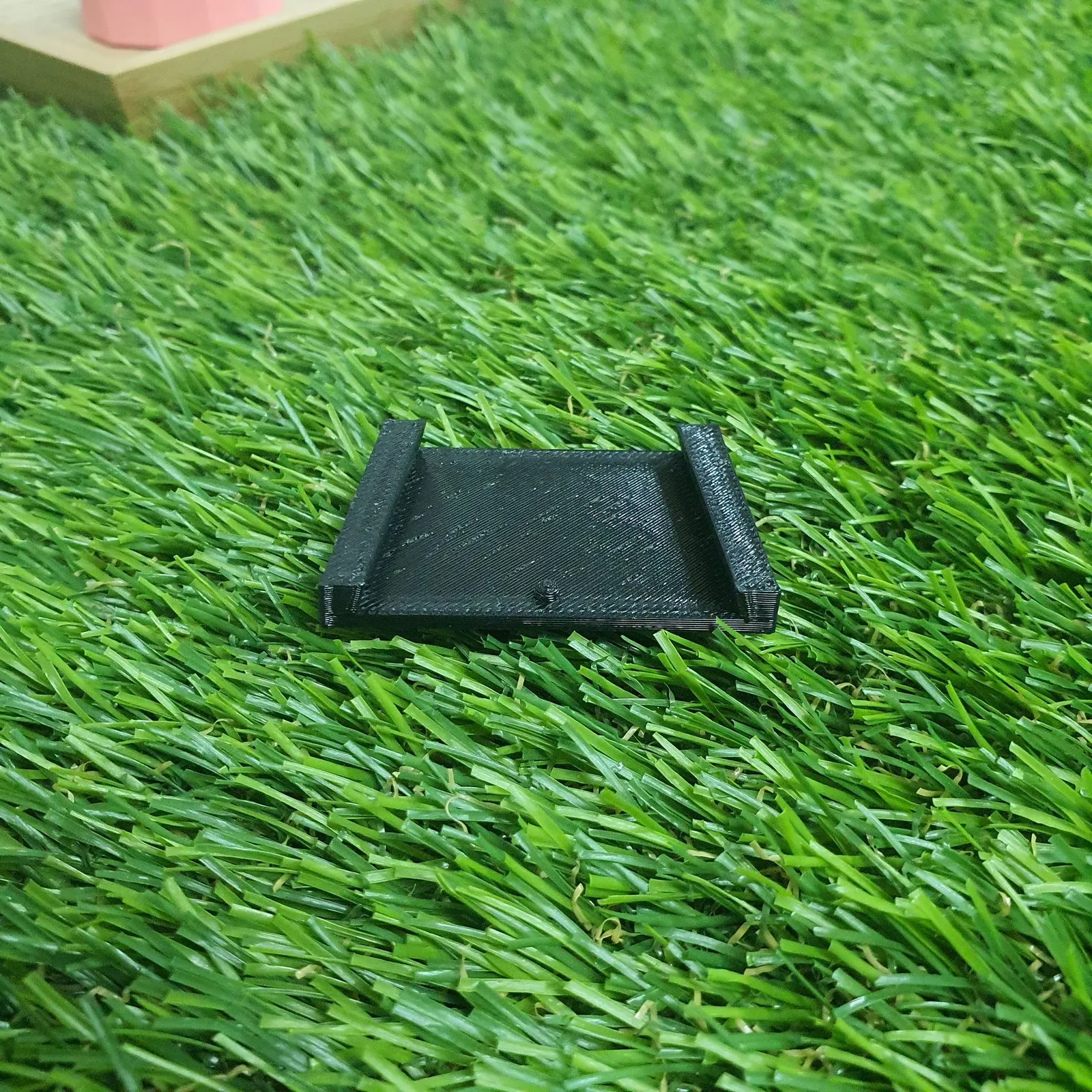 A small black 3D printed tray with a raised lip along two sides and a small peg in the middle, on artificial grass