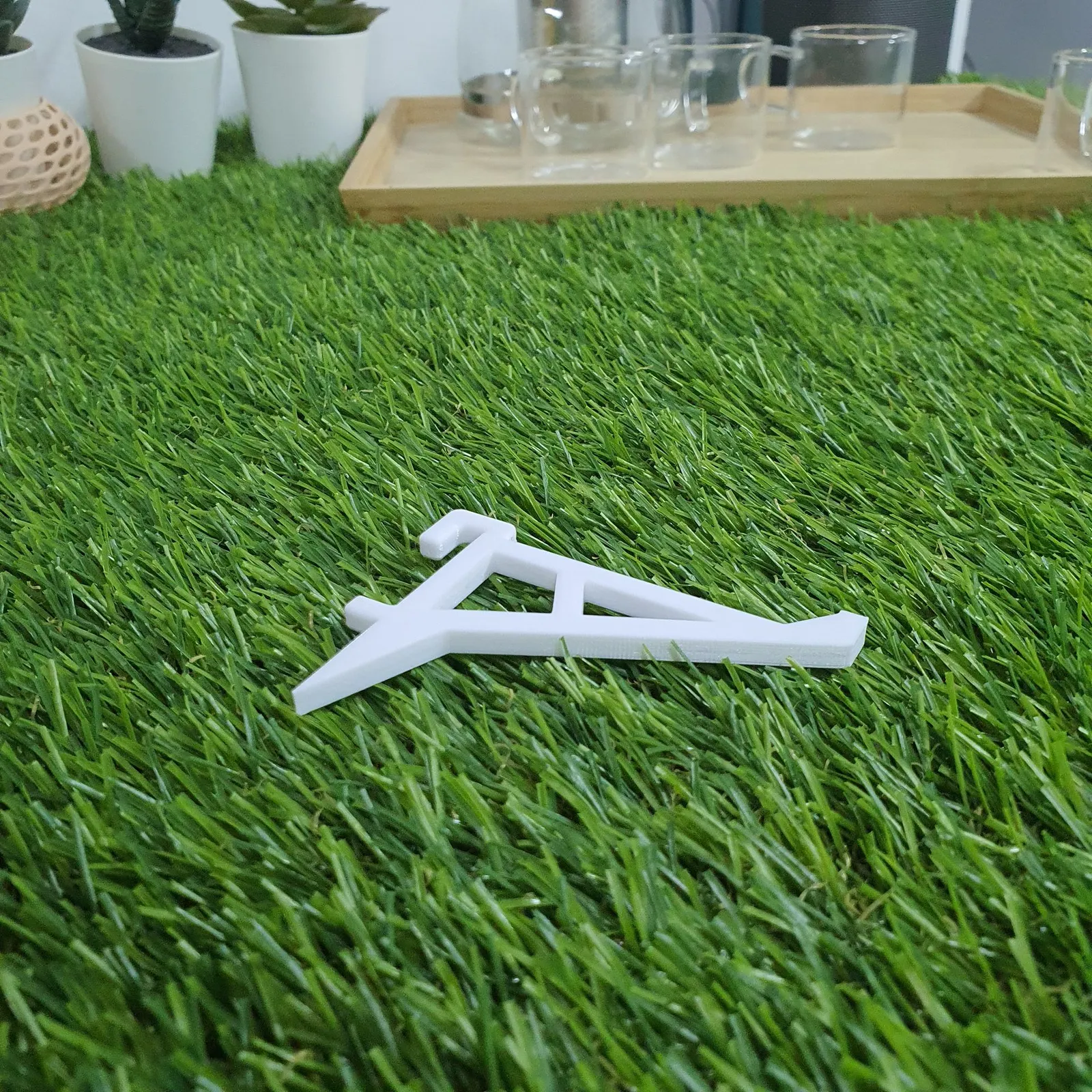 A white 3D printed support arm shaped like a braced triangle, with a hook at the top, lying on artificial grass