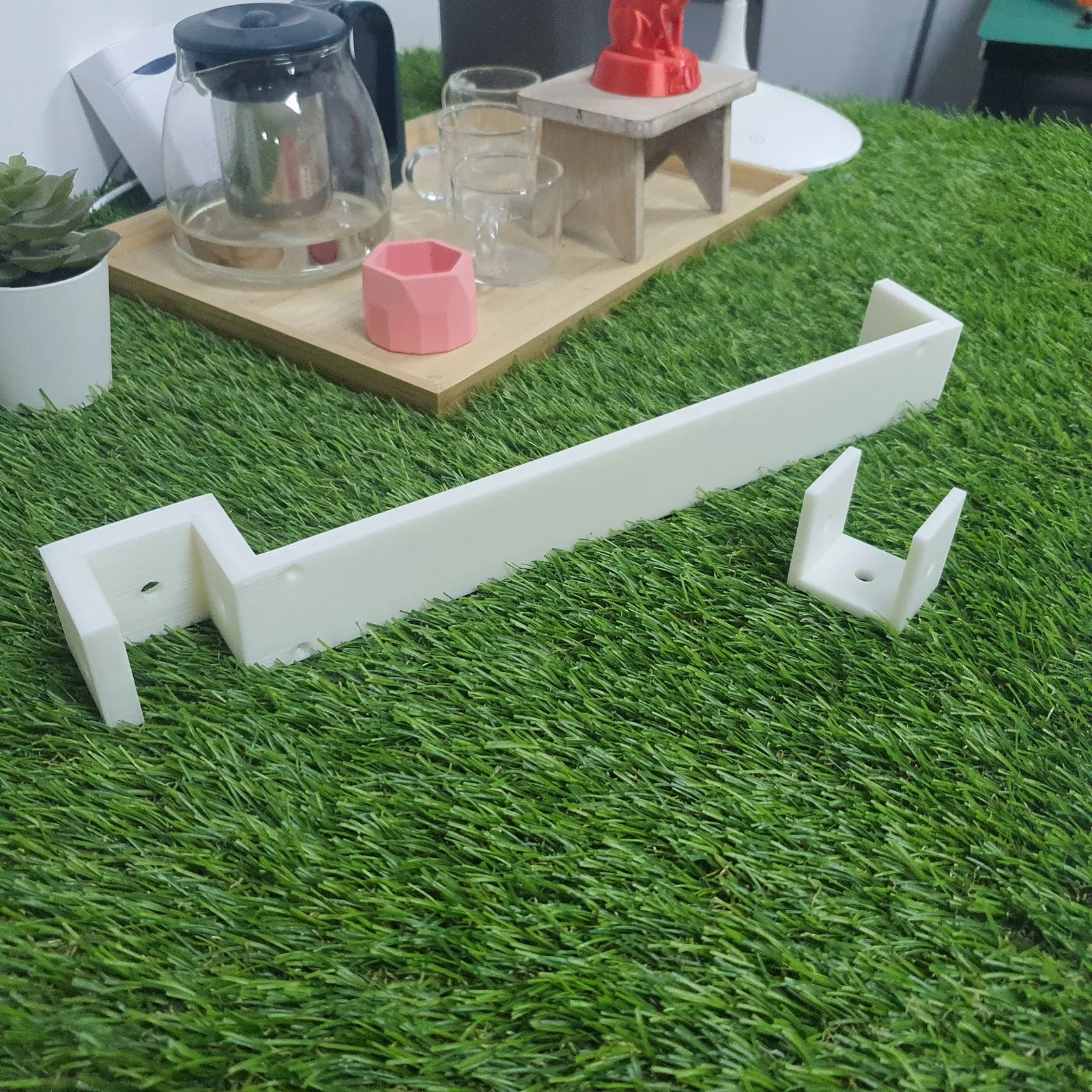 A long white 3D printed wall bracket with screw holes and a bent end, beside a small white U-shaped clip, on artificial grass