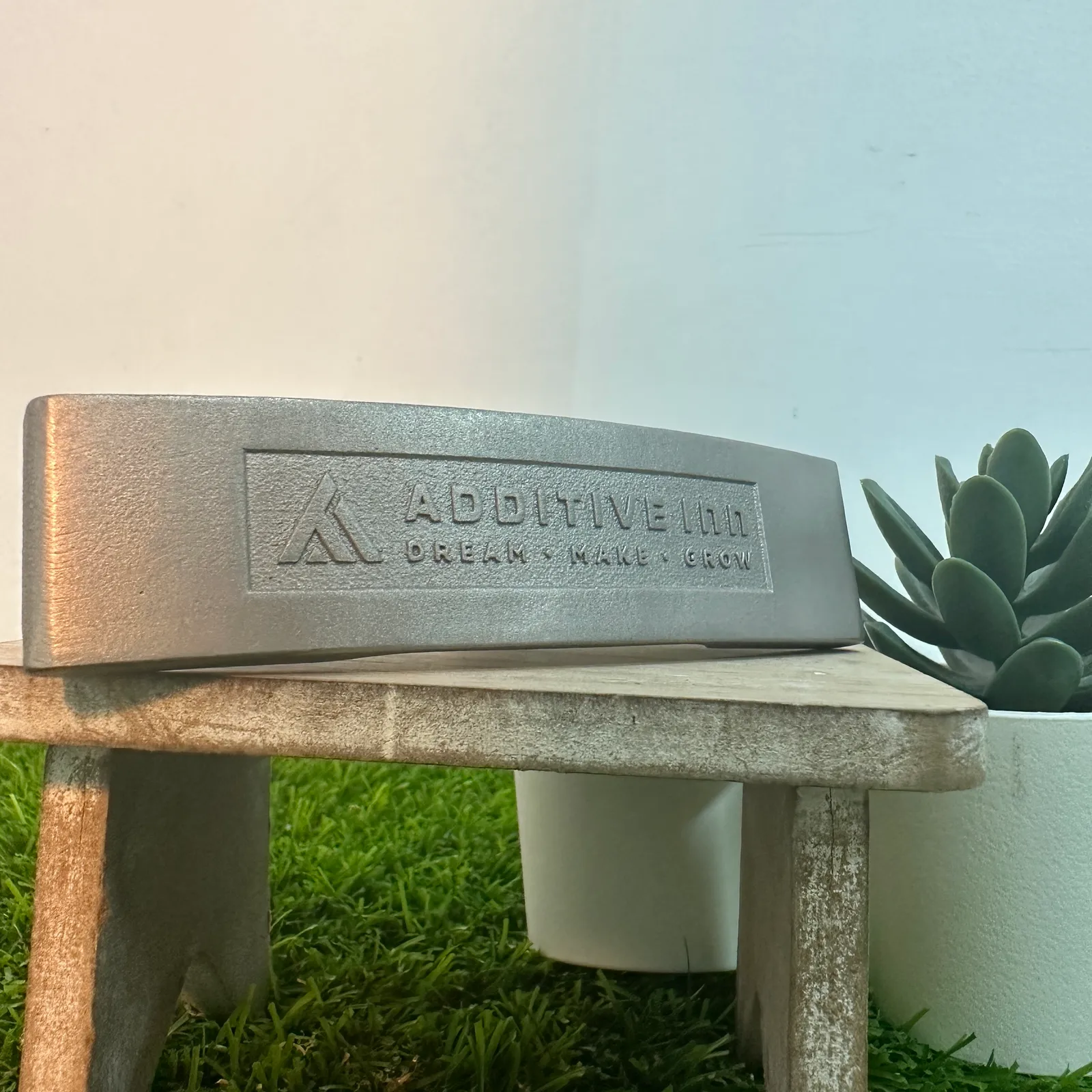 The face of the finished putter head resting on a small wooden stand beside a potted succulent, the engraved logo panel centred on the face