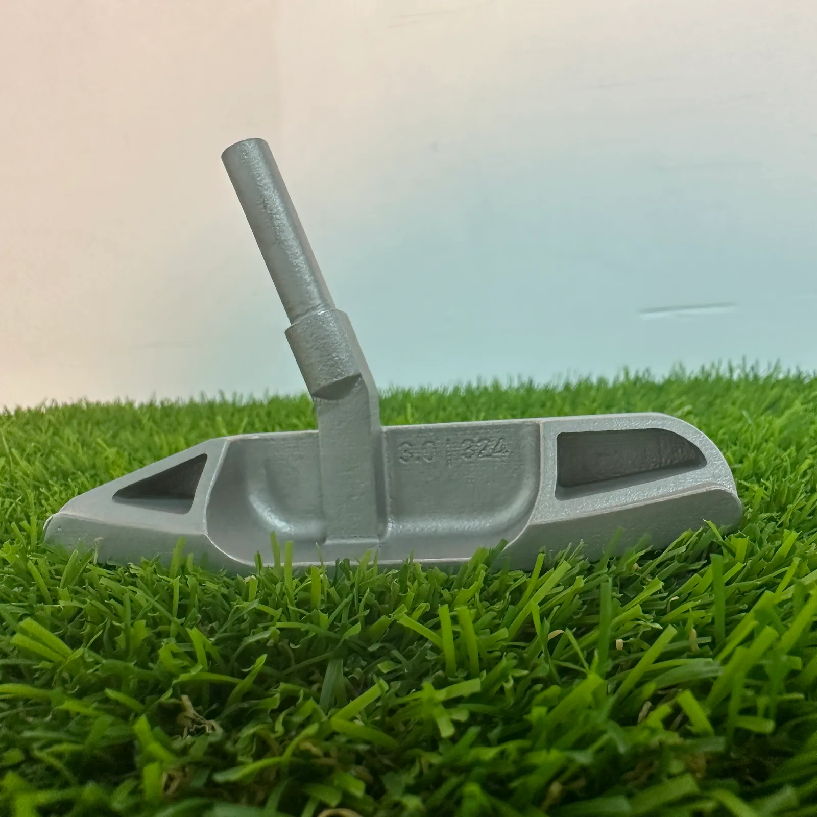 The finished putter head on artificial grass from a back corner, showing the angled hosel post and the triangular heel pocket