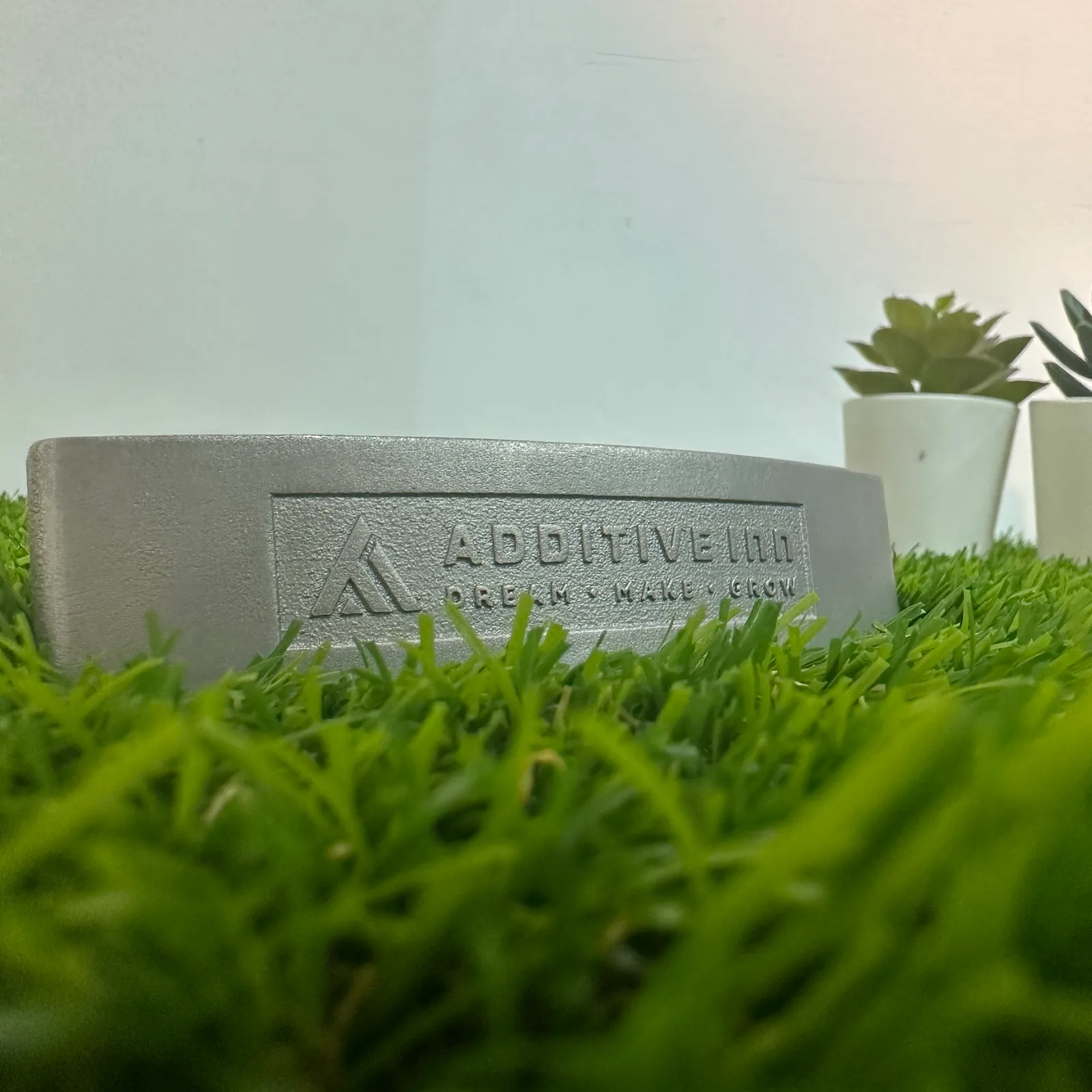 The face of the putter head low on artificial grass, with the Additive Inn logo and Dream Make Grow engraved inside a framed panel