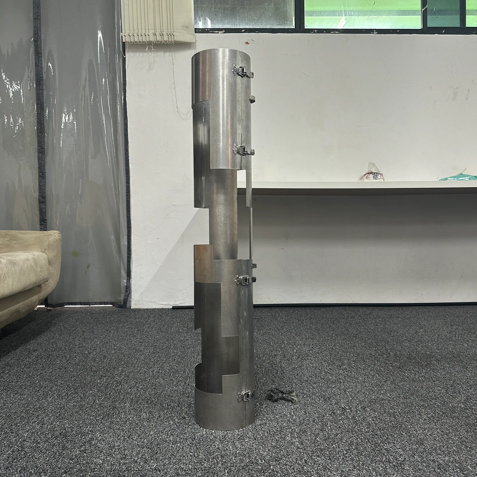 The finished stainless steel sleeve standing upright in the studio, with its windows and slots cut along its length and hinges and latches on its seams