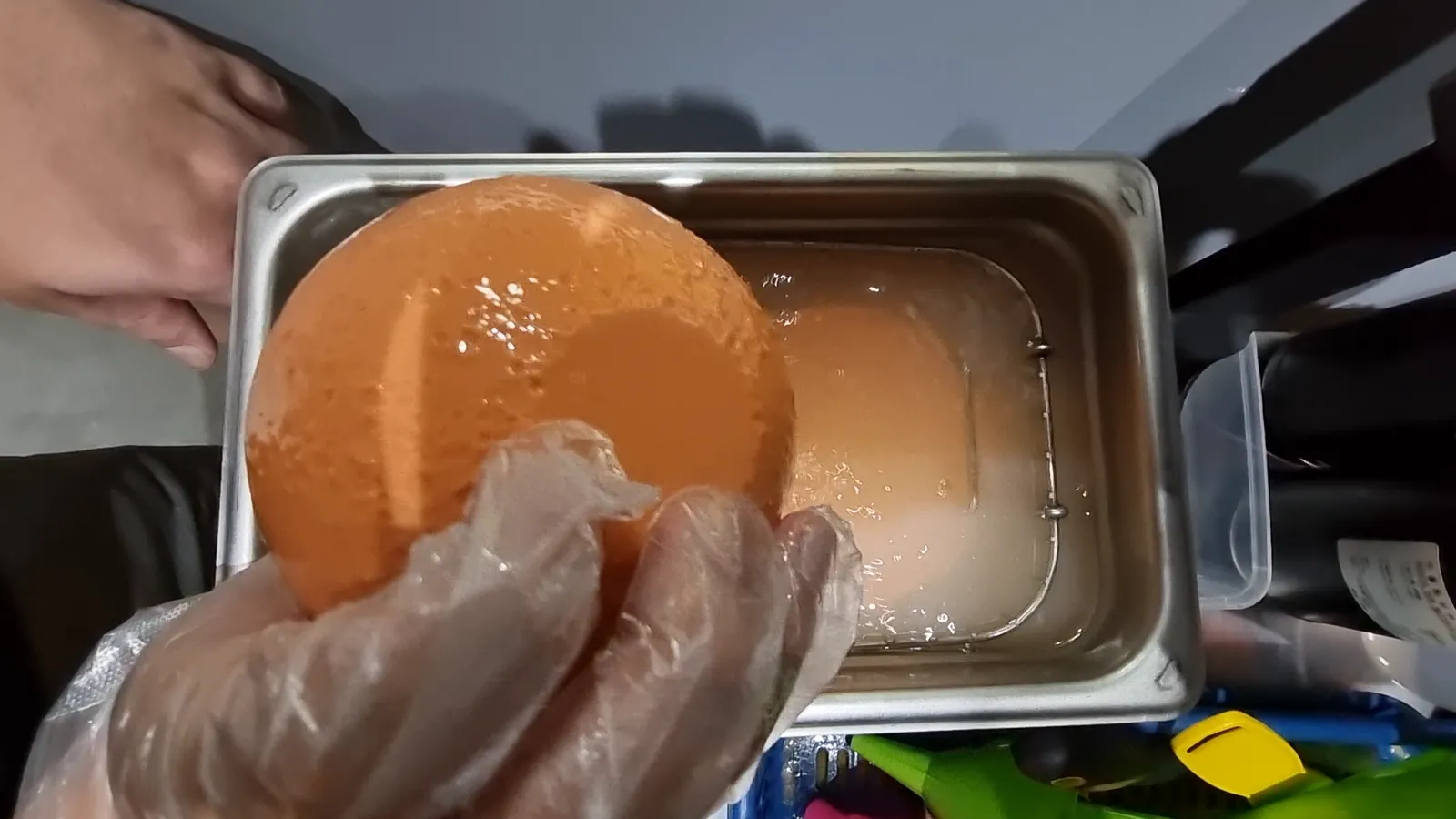 A gloved hand lifting the orange resin print of the moon out of a metal tray of cleaning liquid