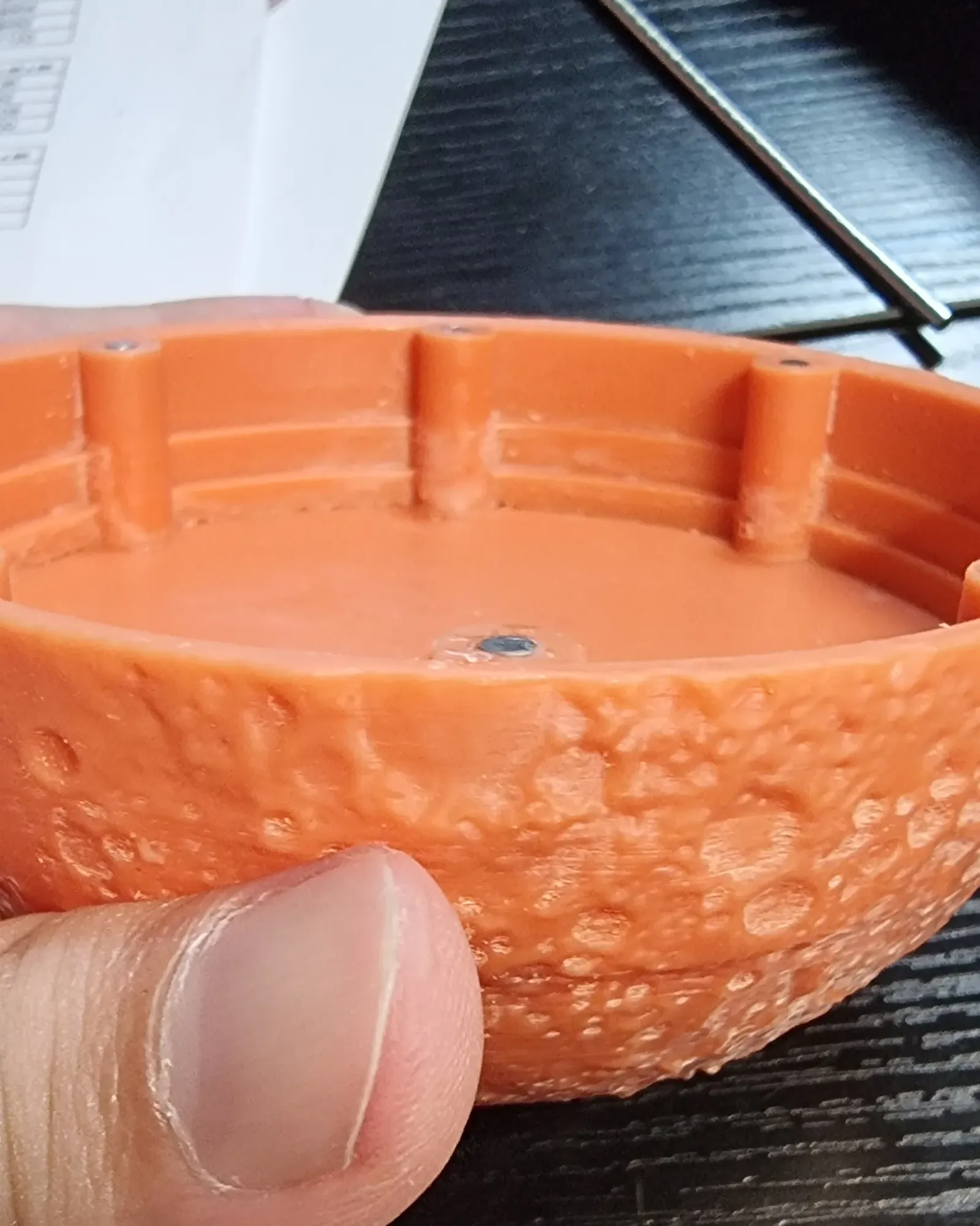 Close-up of one unpainted orange resin half held in a hand, showing the crater relief and the edge of the rim