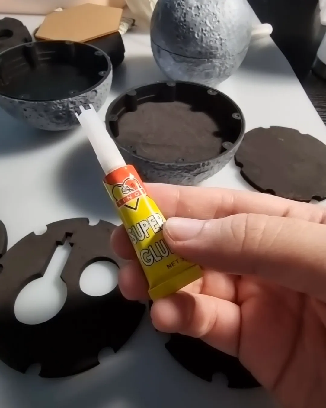 A hand holding a tube of glue over the cut black foam layers, with a painted half of the moon behind
