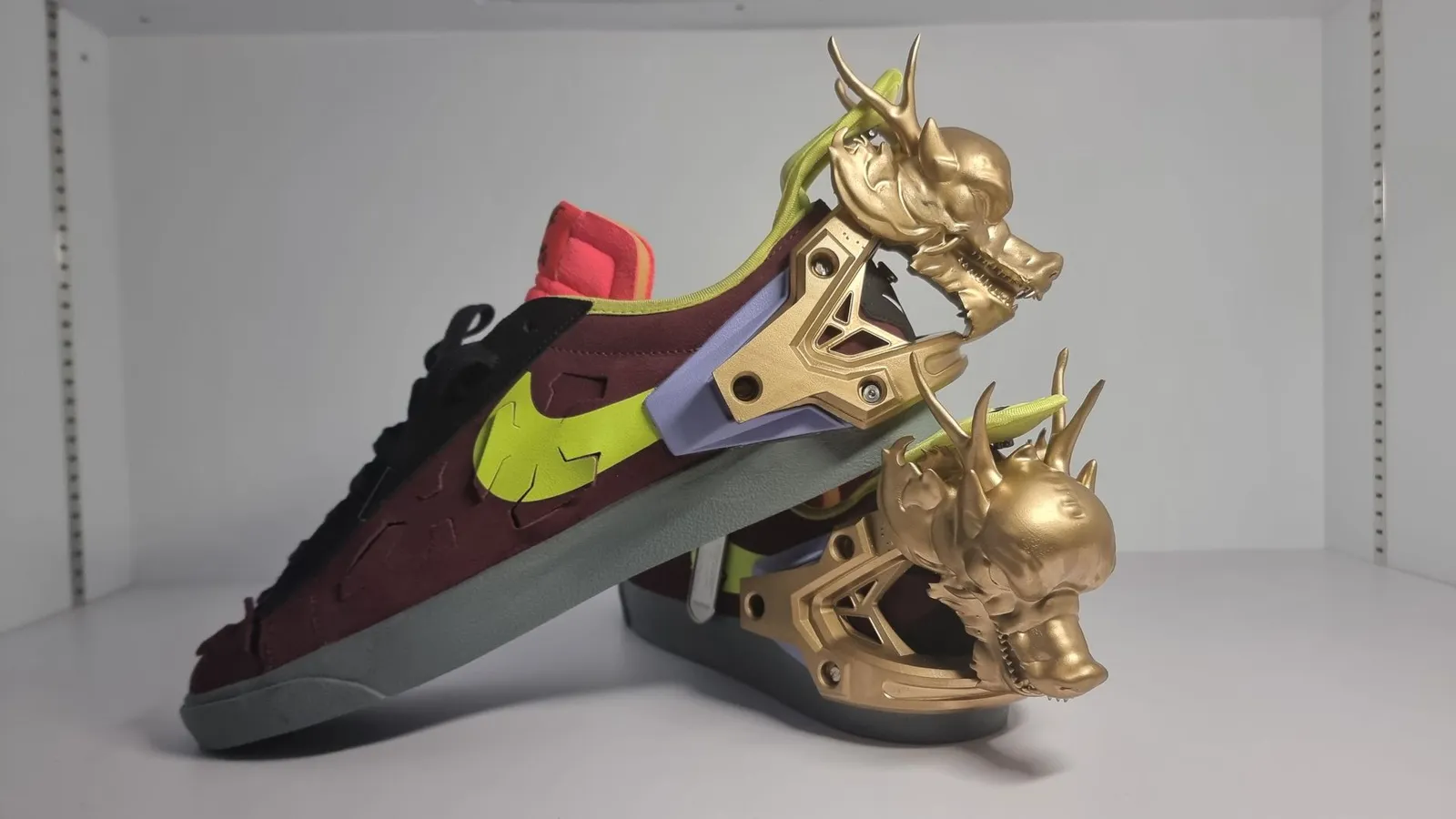 Side view of the pair of trainers on a white shelf, a gold dragon fixed over each heel by screws through its bracket