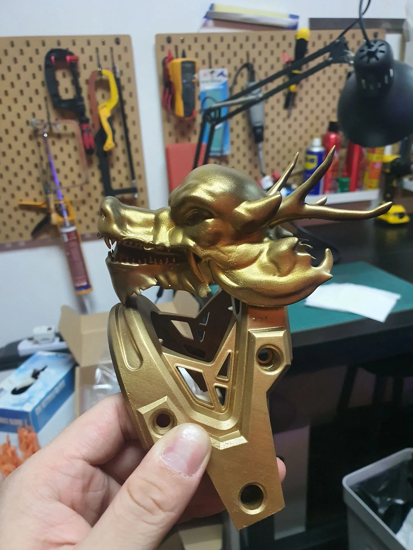 A hand holding one dragon head and bracket after gold spray paint, in the workshop with tools on a pegboard behind