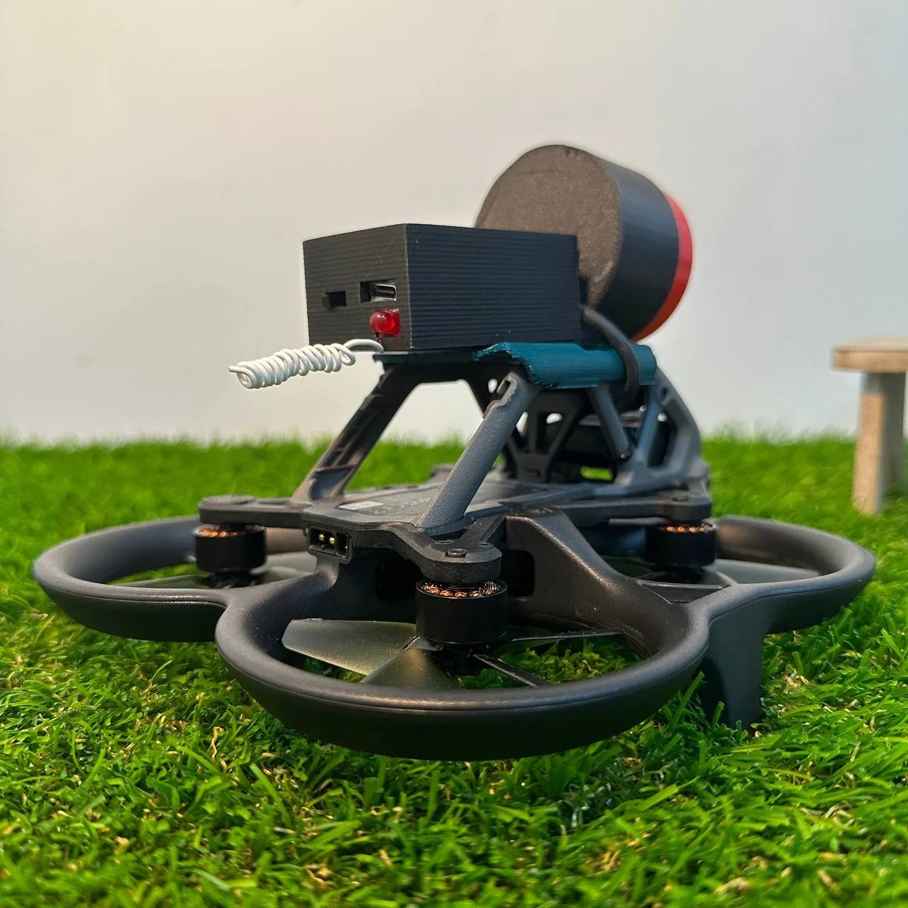 The drone from behind, showing the black power box with its ports and a red indicator light, the spotlight behind it on the teal mount