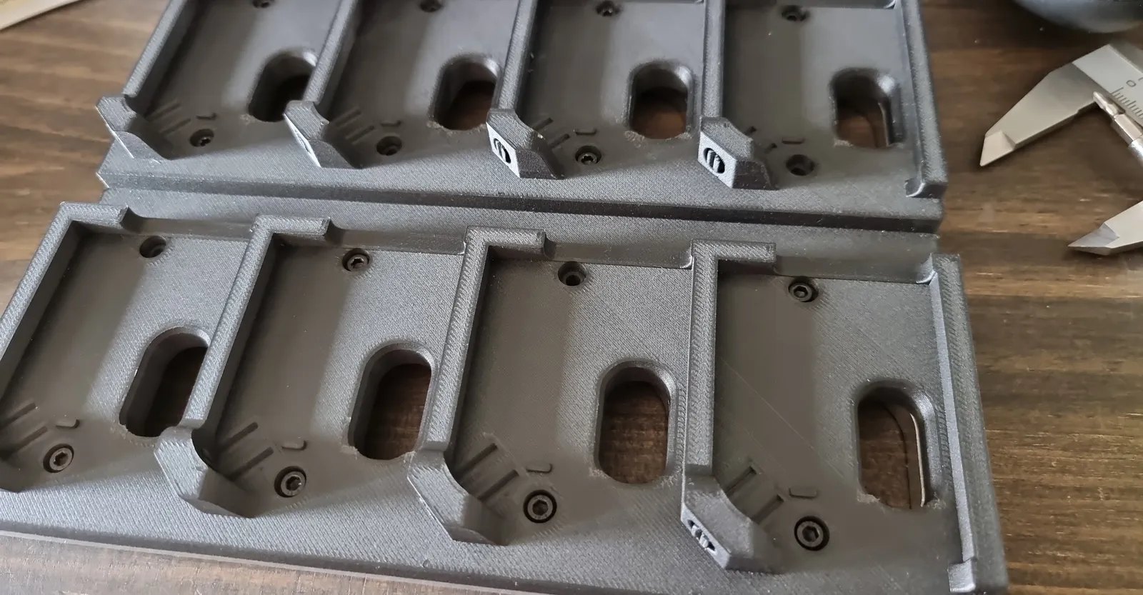 Two dark grey printed trays of four pockets each, with an oval window and screw holes in every pocket, on a wooden desk beside a vernier calliper