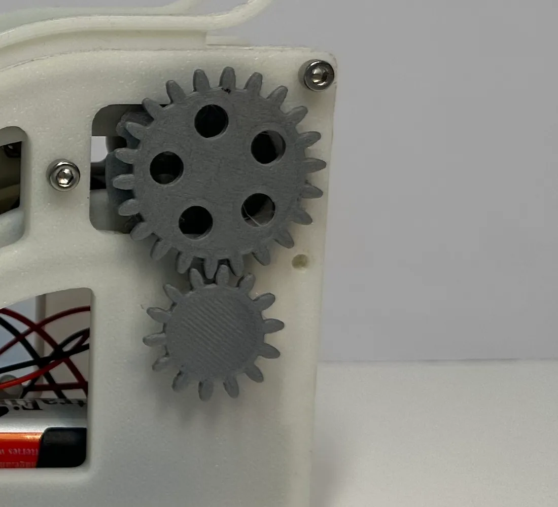 Close-up of two grey printed spur gears meshing on the white side frame, with red and black wiring and the battery visible through a cut-out