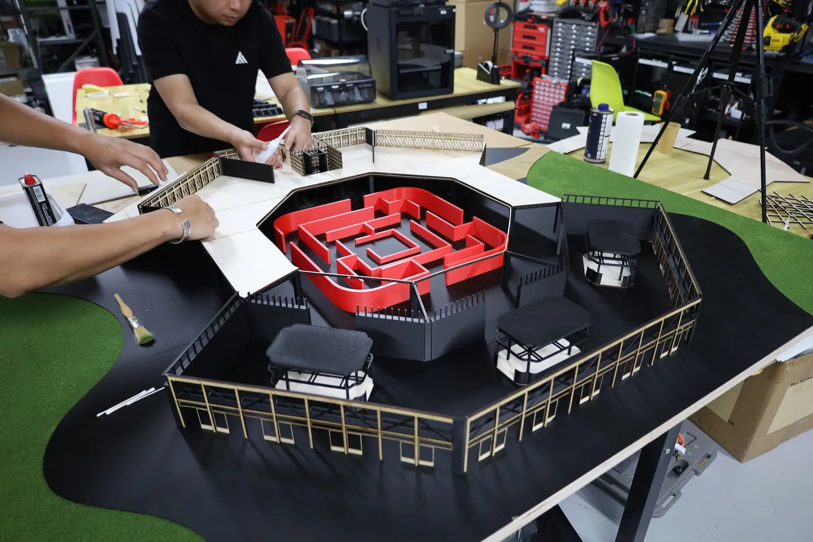 Two of the team gluing plywood upper-level slabs and lattice rails onto the model, with the red maze in the octagonal courtyard below