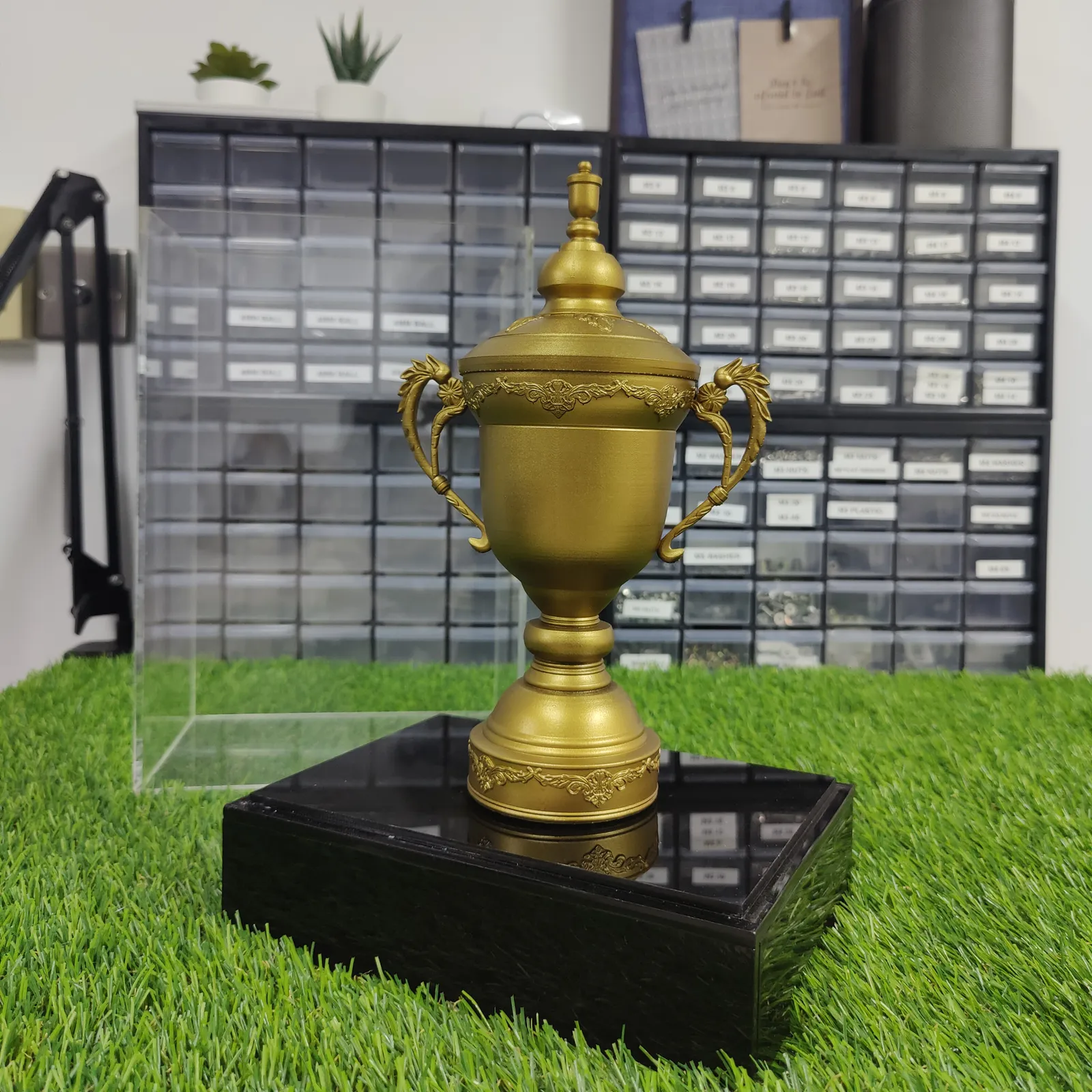 The finished Congress Cup replica in matte gold, with scrolled handles and a domed lid, on a glossy black base beside its clear acrylic case