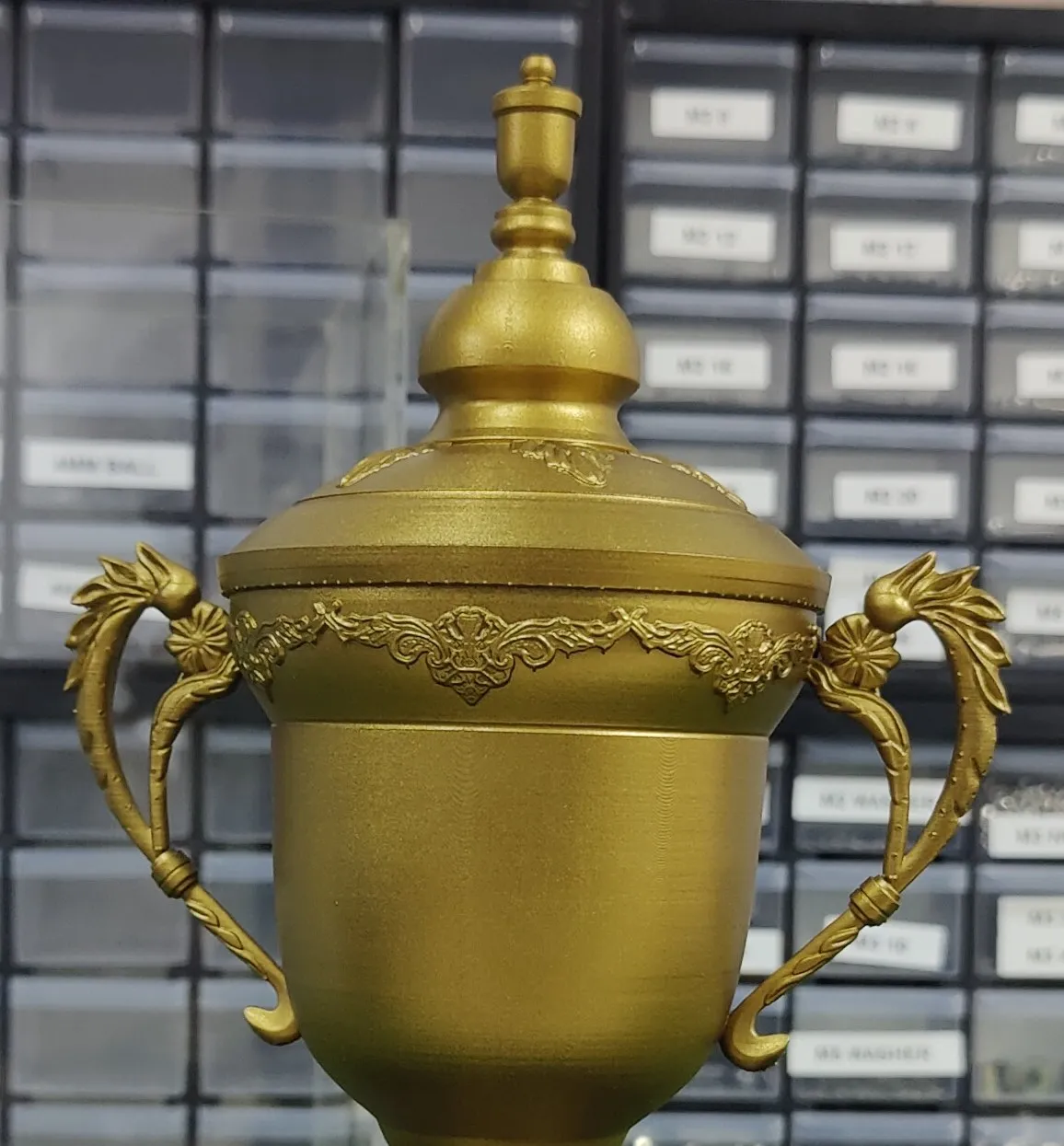 Close-up of the replica's domed lid and finial, the band of leaf ornament below it and the two scrolled handles, all in matte gold