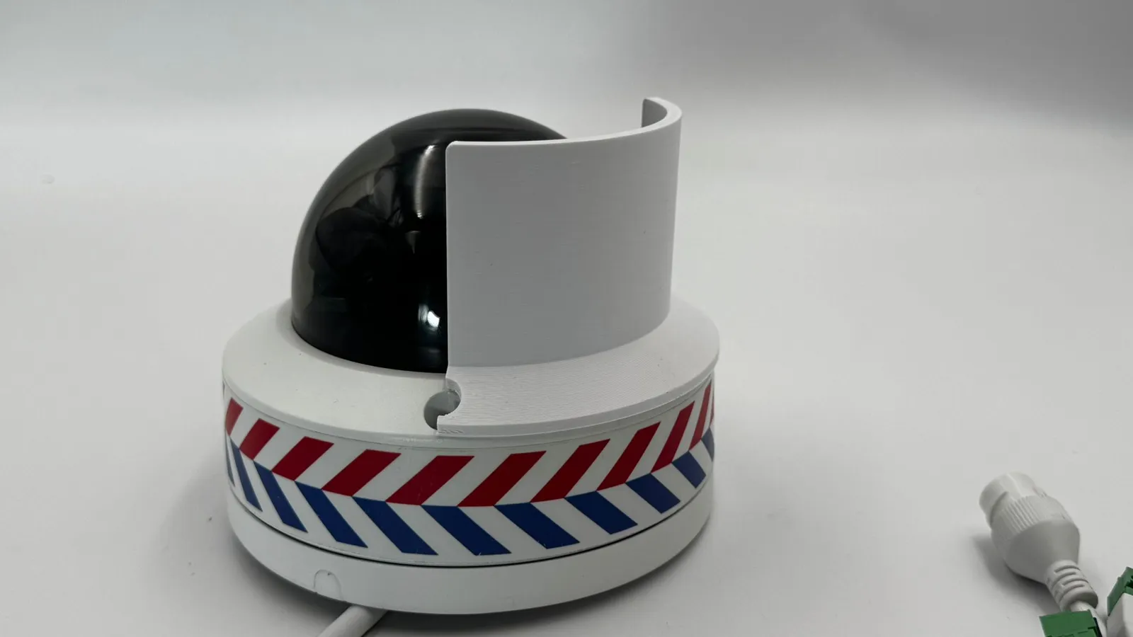 A white 3D printed privacy shield standing beside the black dome of a security camera, its foot notched around one of the camera's screw holes