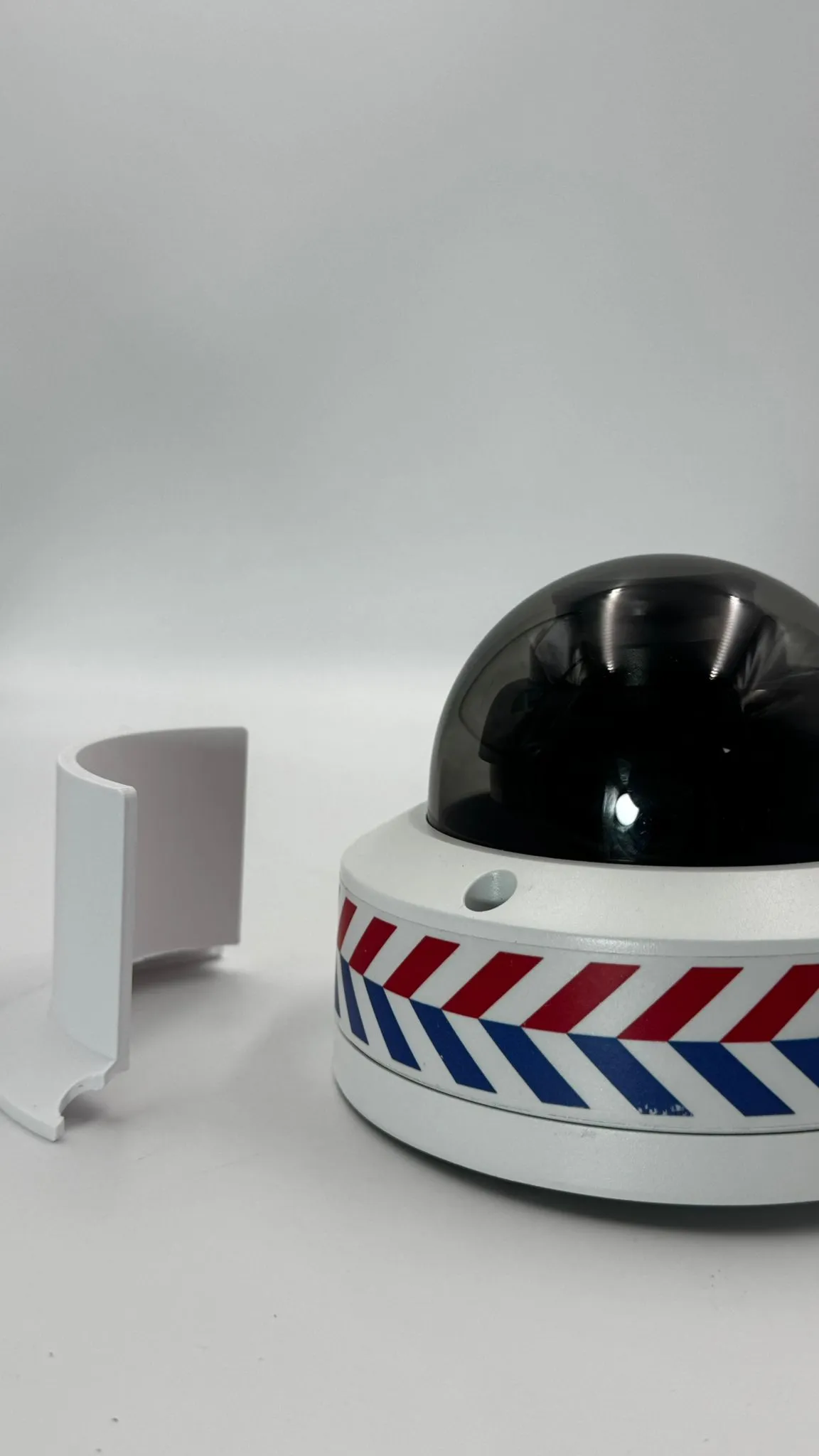 A printed white shield segment standing on its own beside the dome camera, showing its curved wall and notched foot