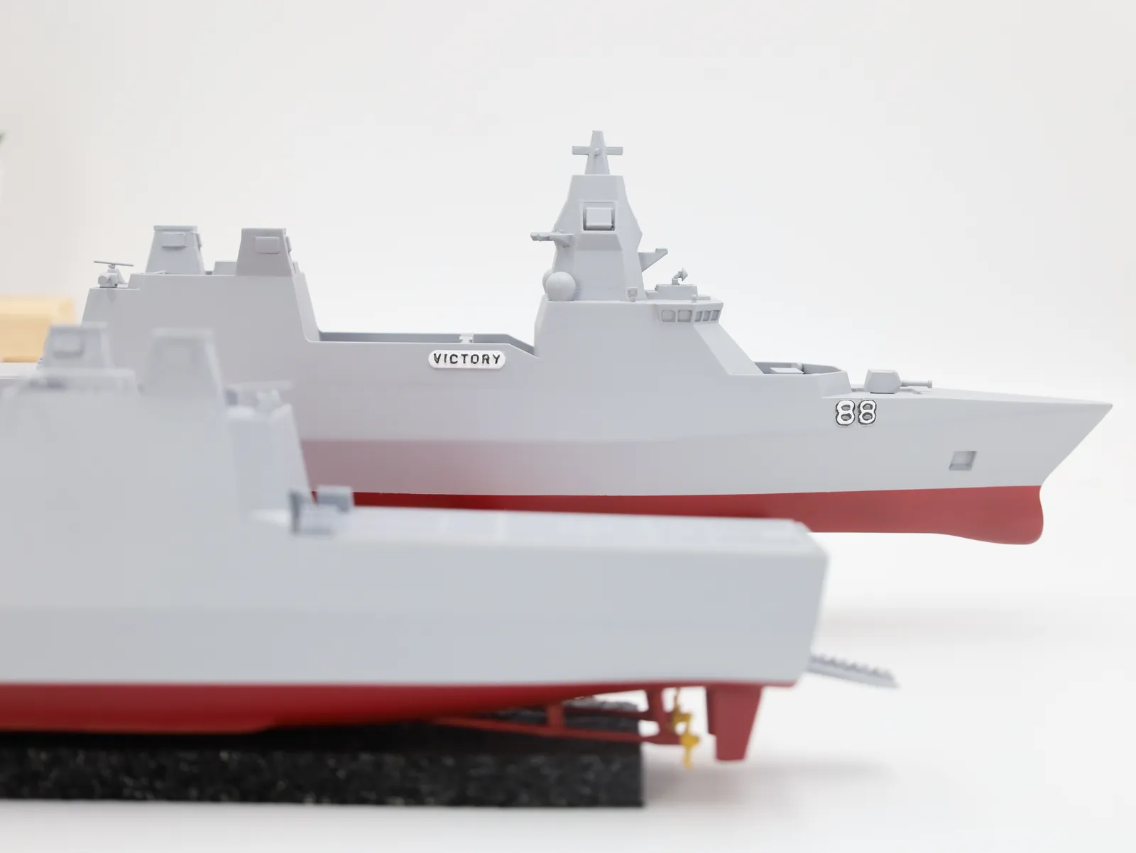 Two finished frigate models in a bright studio: one seen side-on with VICTORY and 88 on its hull, the stern of the second in front with its rudders and ramp
