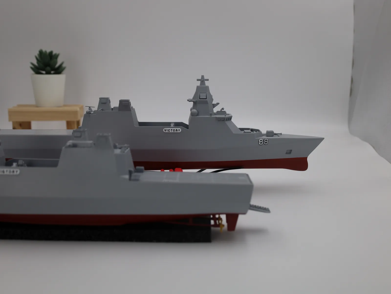 Two finished VICTORY frigate models one behind the other on black stands, grey with red lower hulls, a small plant behind them