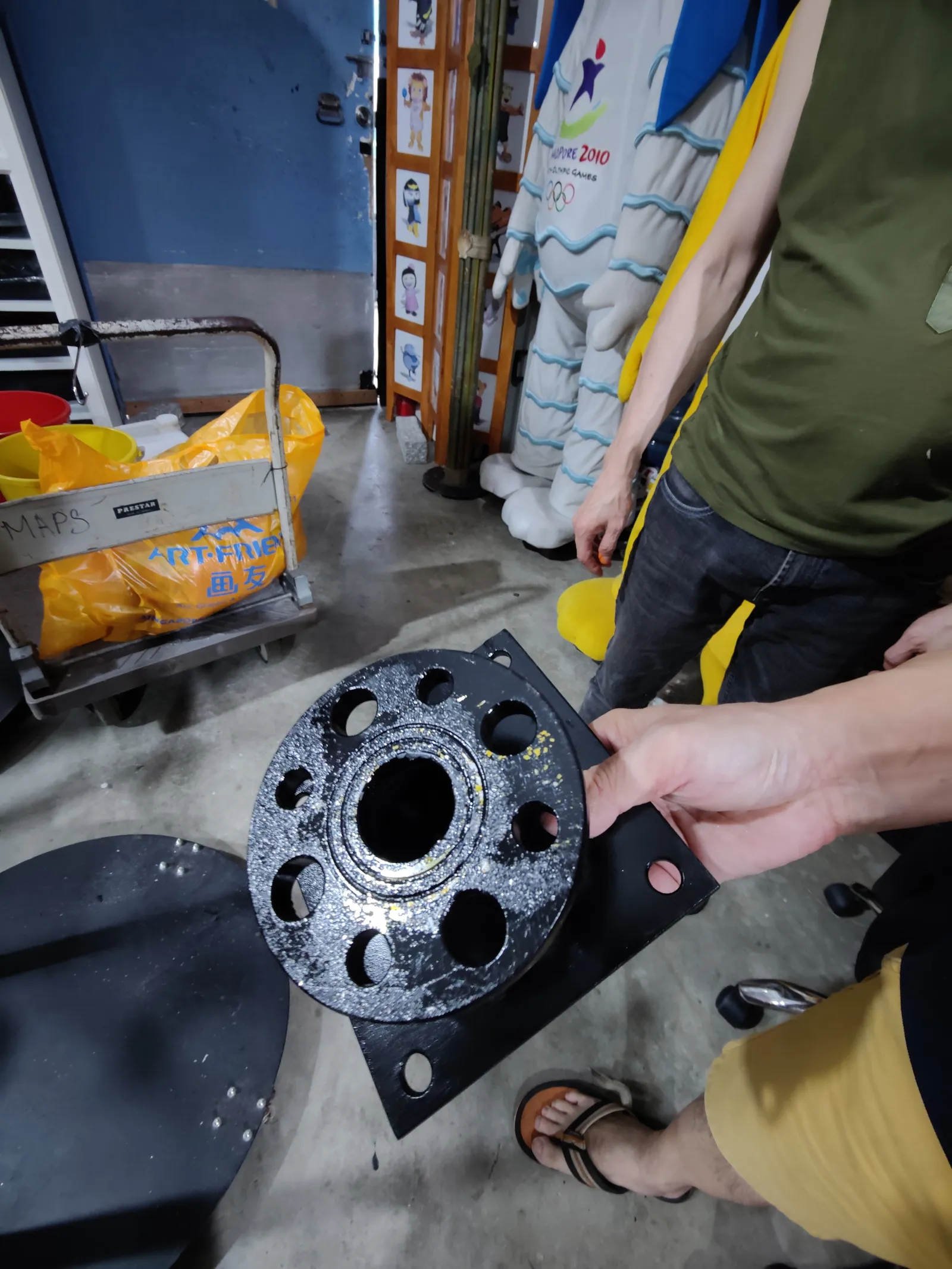 A hand holding one of the black metal wall brackets: a square base plate with a drilled round flange