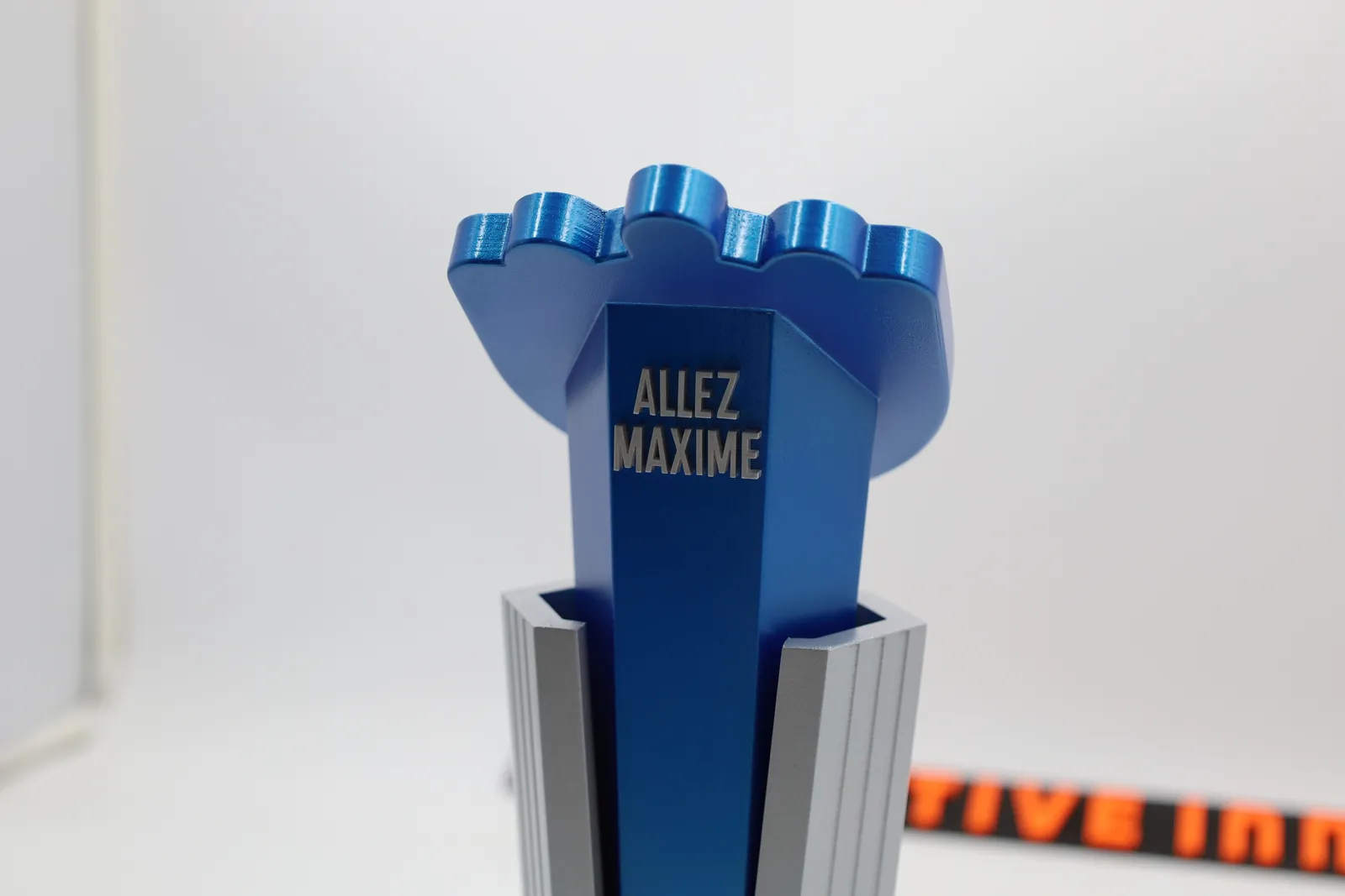 Close-up of the back of the glove, its fingertips in bright blue, with ALLEZ MAXIME lettered on the stem above the silver pillar