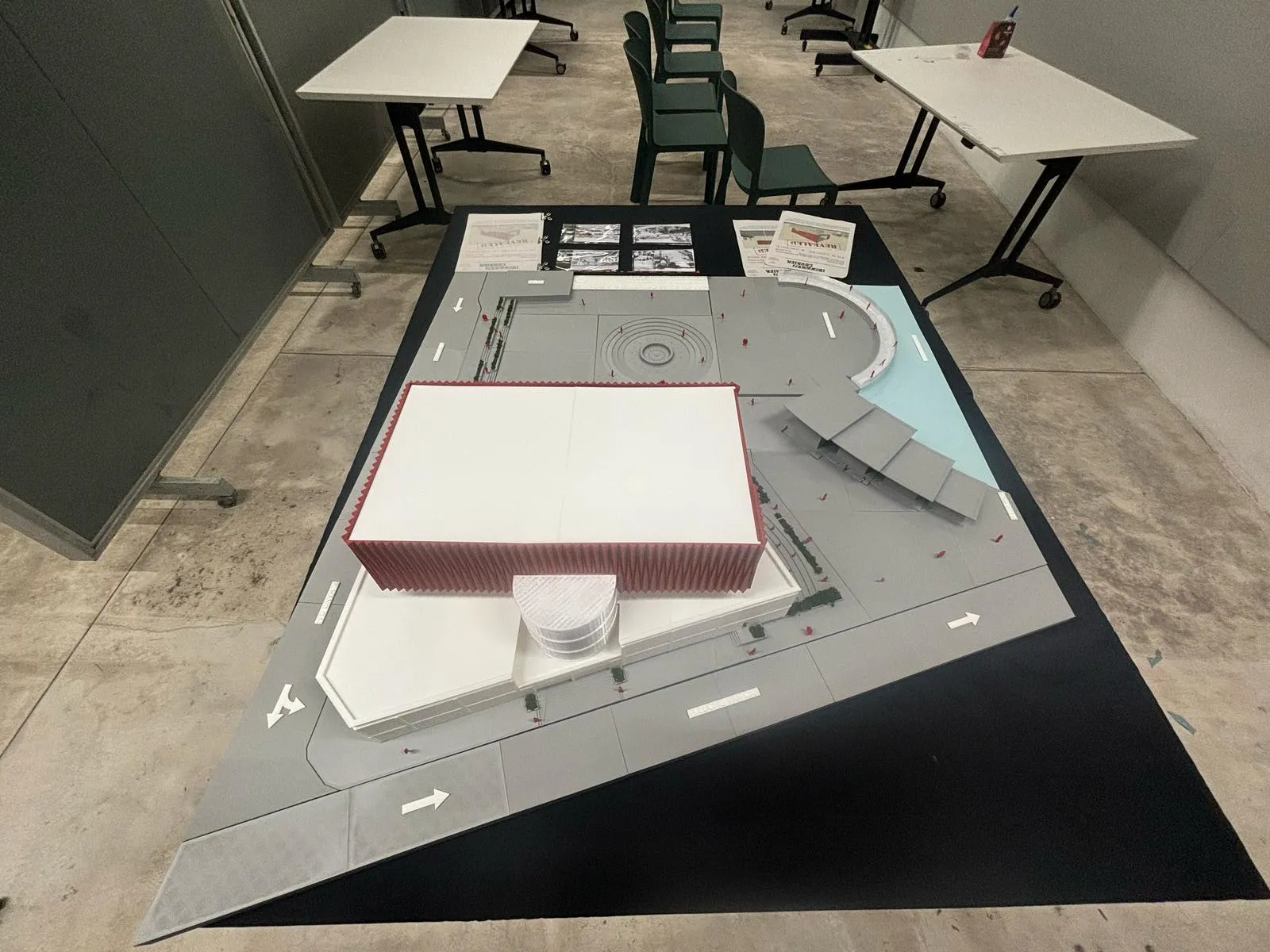 The model from above at an angle: the red fluted block over the white building, grey site panels, road arrows and a blue lakeside edge