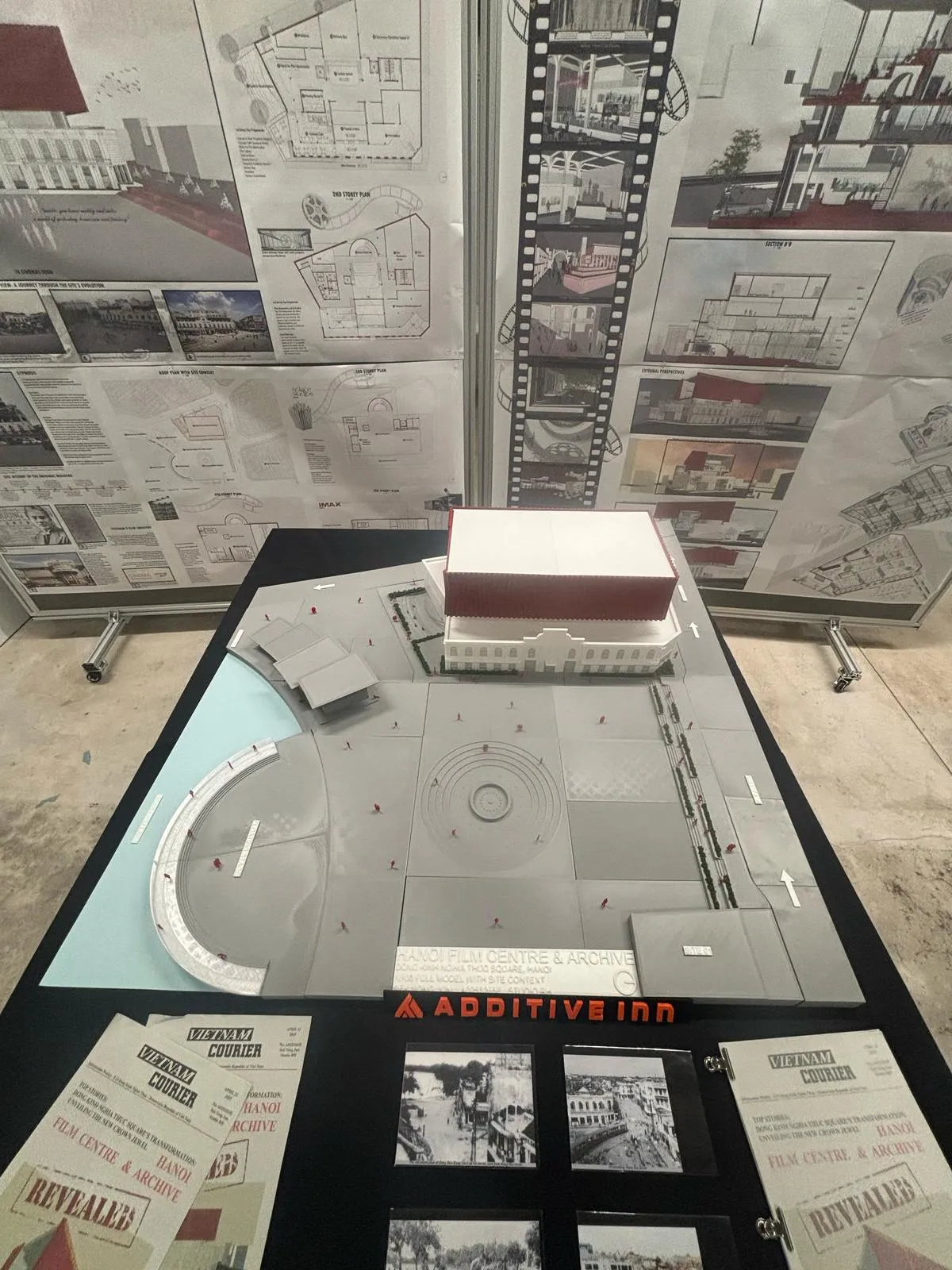 Looking down on the whole site: the building at the back, a round stepped plaza in the square, a curved edge along blue water and hedges along the street