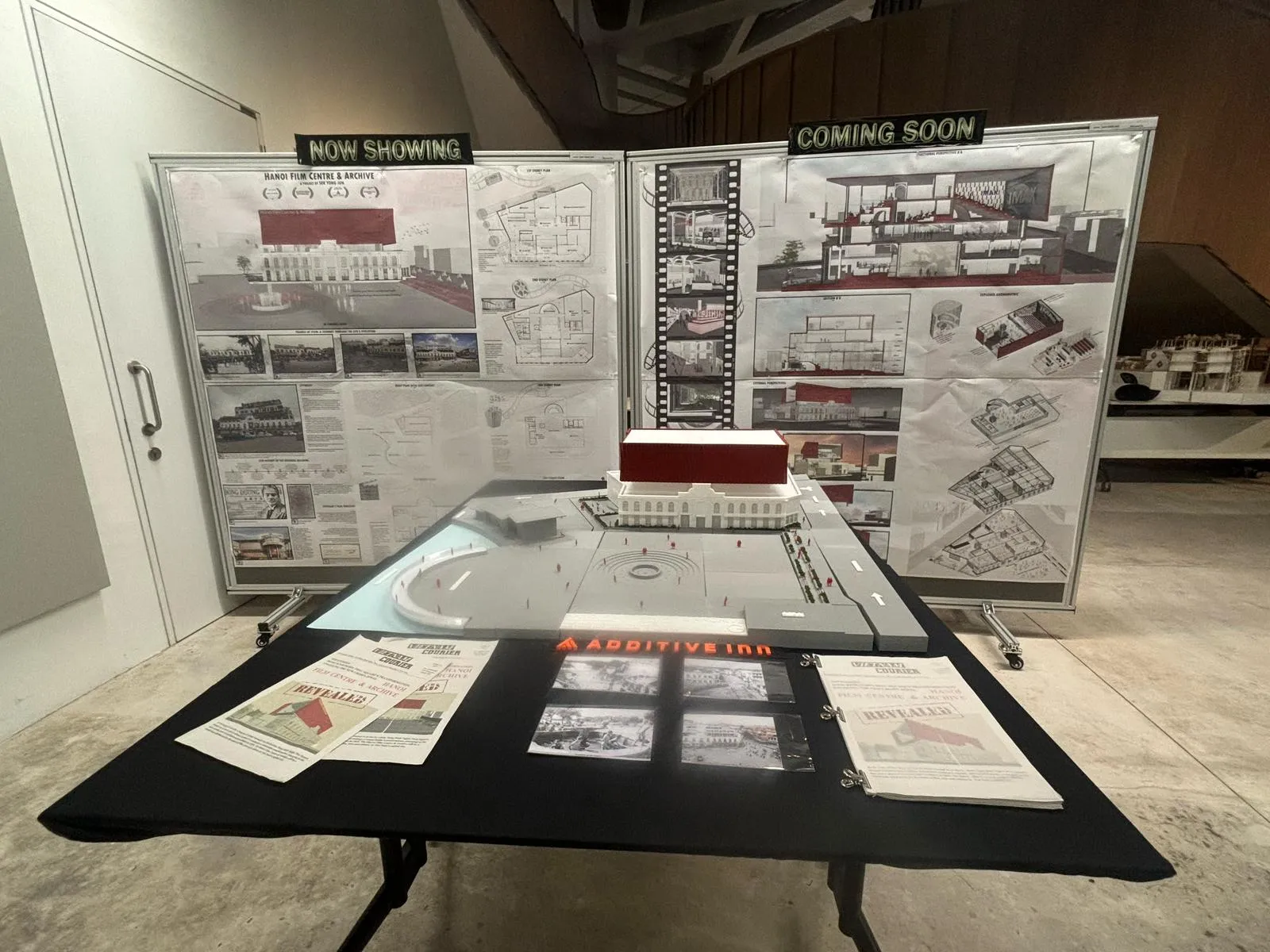 The model on a black table in front of the presentation boards, with printed newspapers and photos laid beside it