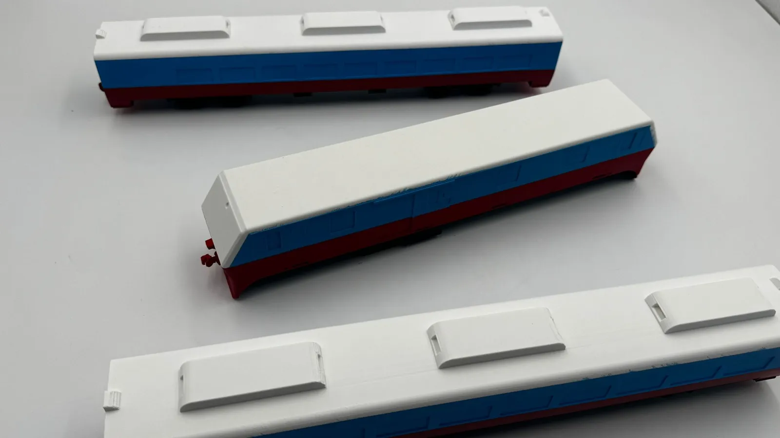 Three model train cars on a white table, white roofs with raised vents above blue sides and red underframes