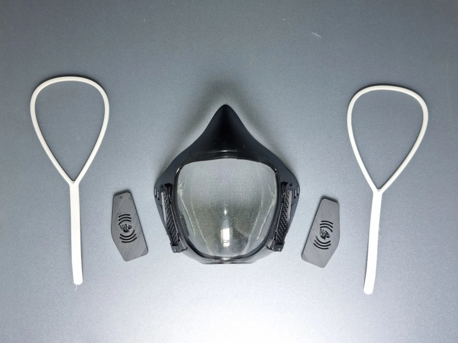 The finished black mask with its clear visor, laid between its two black vent covers and two white ear loops on a grey surface