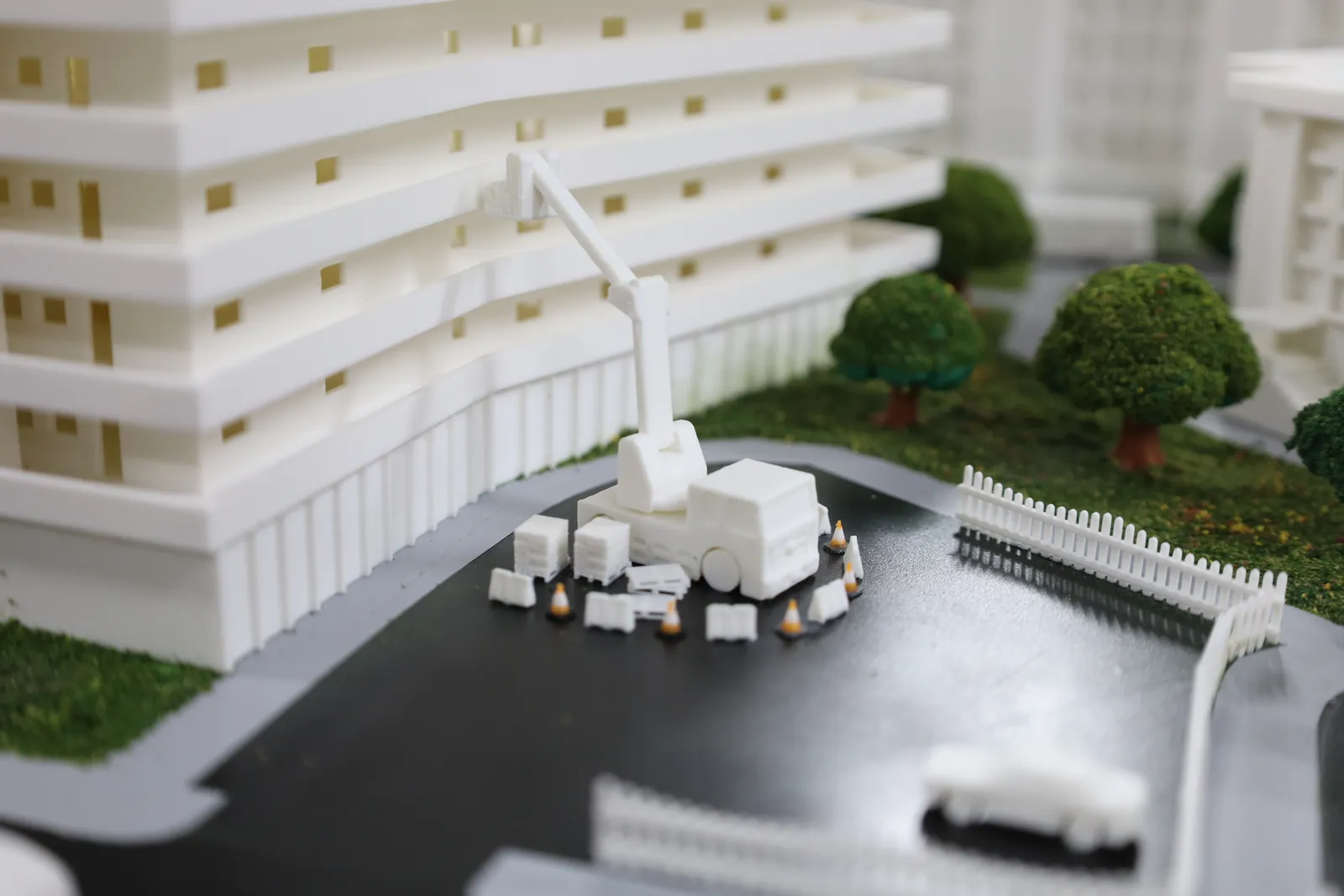 A mobile crane on a paved area beside a curved residential block, ringed by tiny cones and barriers, with a white fence behind