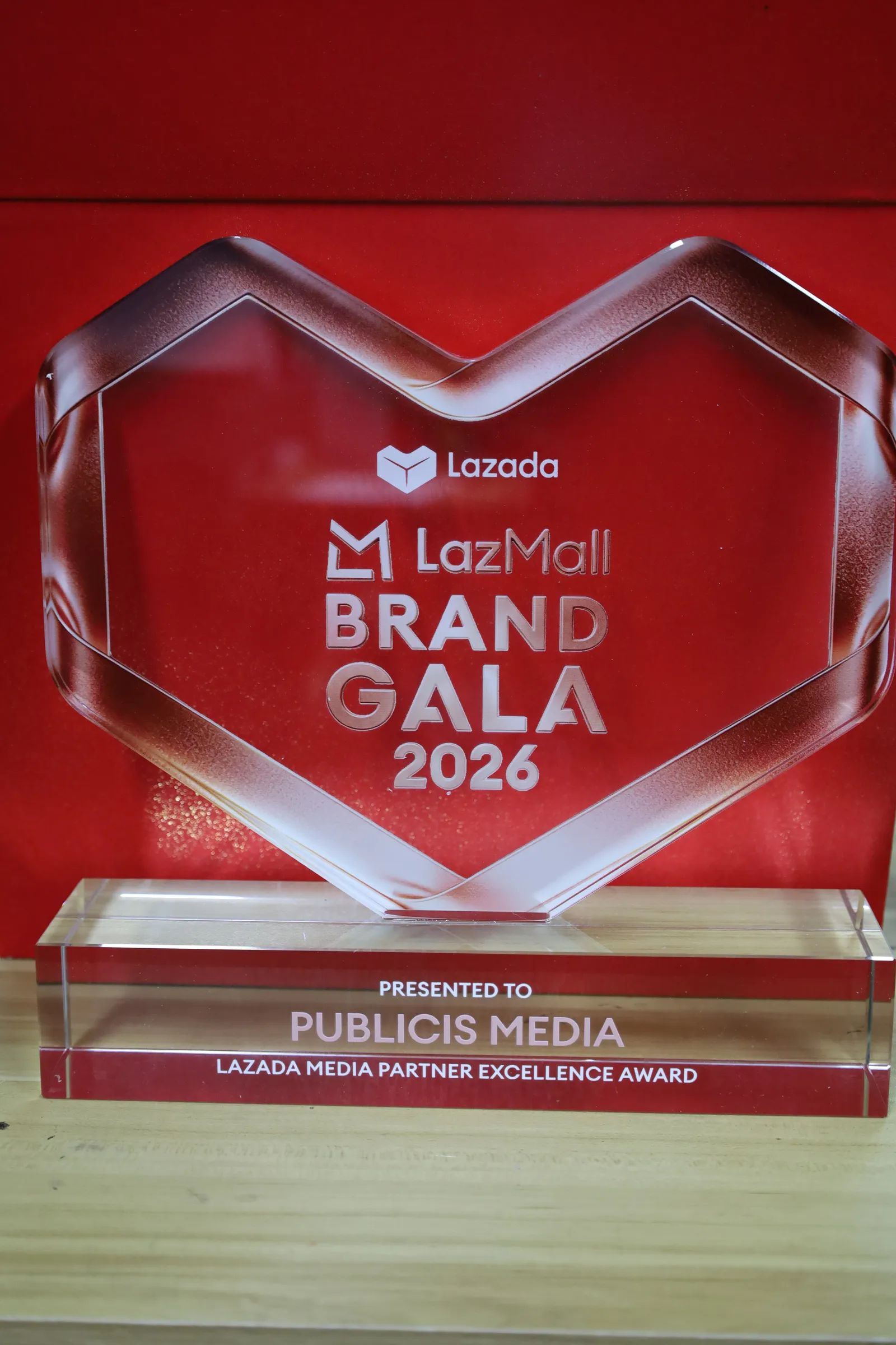 A finished LazMall Brand Gala 2026 heart award in clear acrylic with a rose-red band, presented to Publicis Media, against a red background