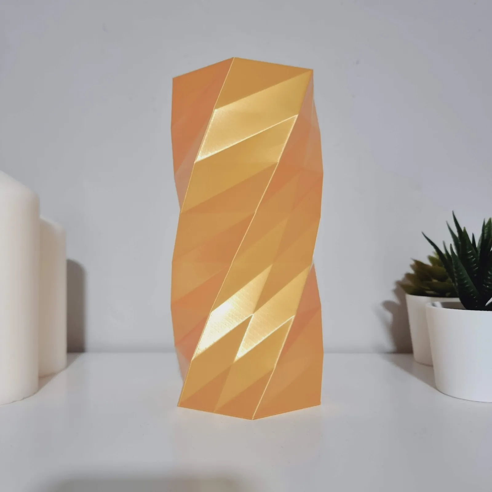 The translucent orange faceted column standing unlit on a white shelf in daylight, beside a candle and a potted plant