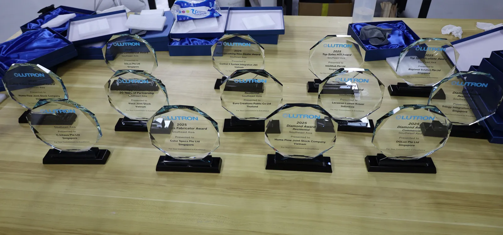 Thirteen faceted glass Lutron awards on black bases laid out on a workbench, with open blue satin-lined boxes behind them