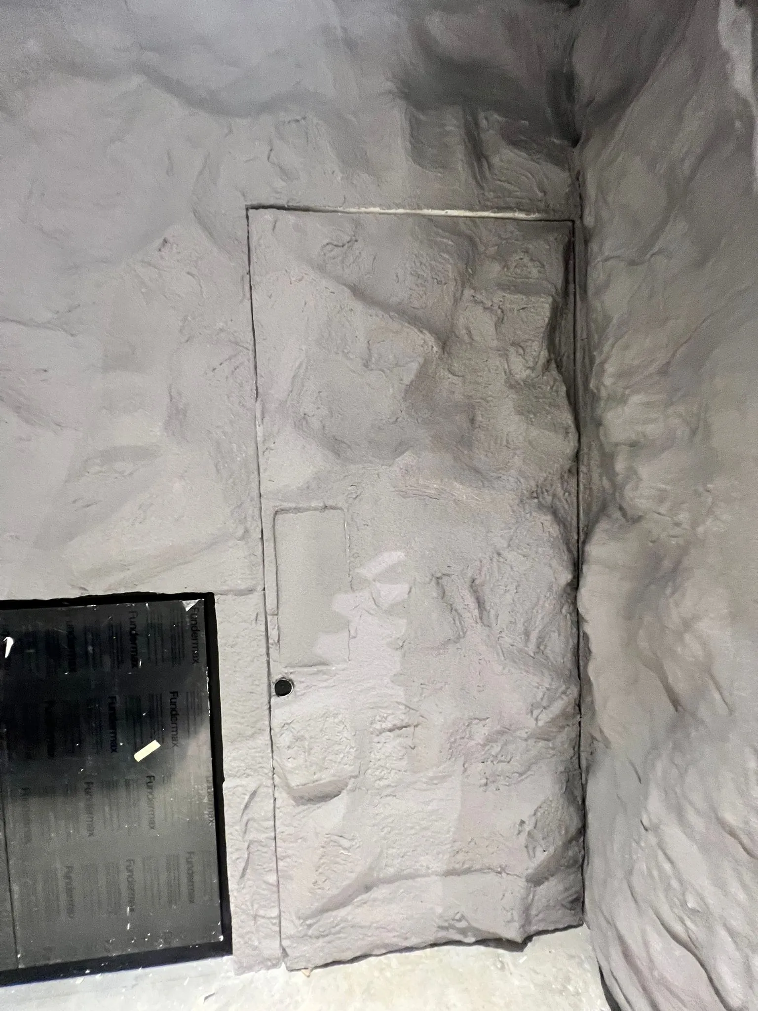 A door clad in the same rock texture as the wall, its outline only just visible beside a dark panel
