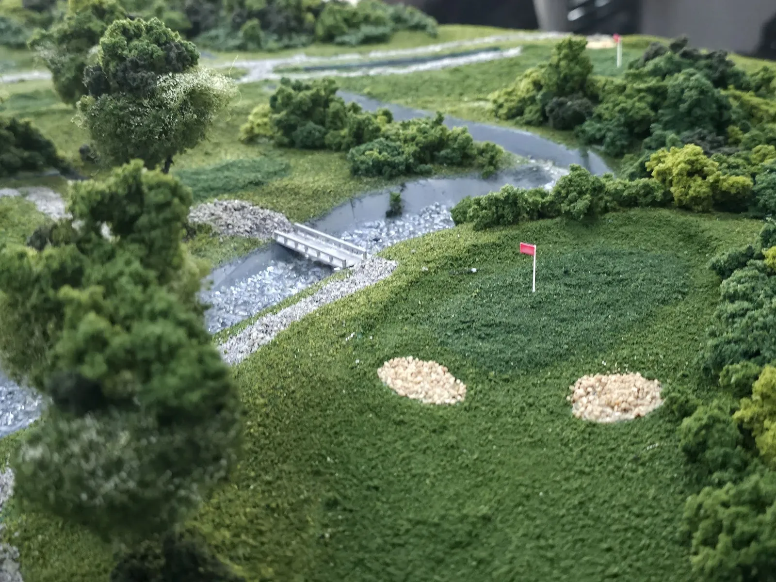 Close-up of a green with a red pin flag and two sand bunkers, a small bridge crossing a drain behind it and trees around the edge